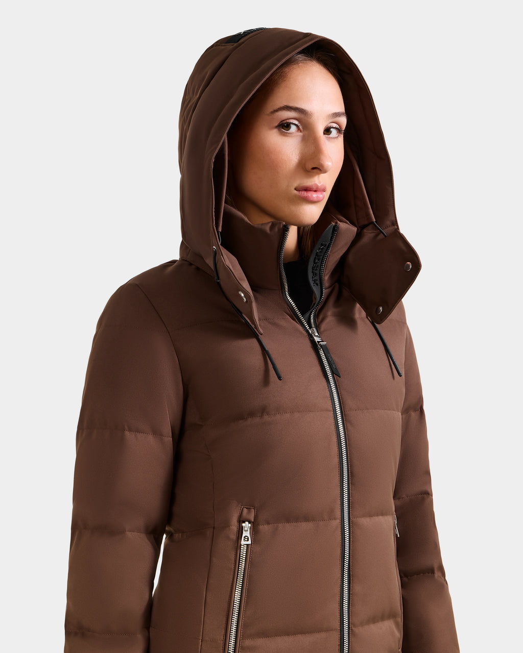 A woman wears the Rudsak MIRIAM - L123544 Mocha down puffer jacket, featuring a brown weather-resistant shell and zippered pockets, with the hood up, looking slightly to the side against a light gray background.
