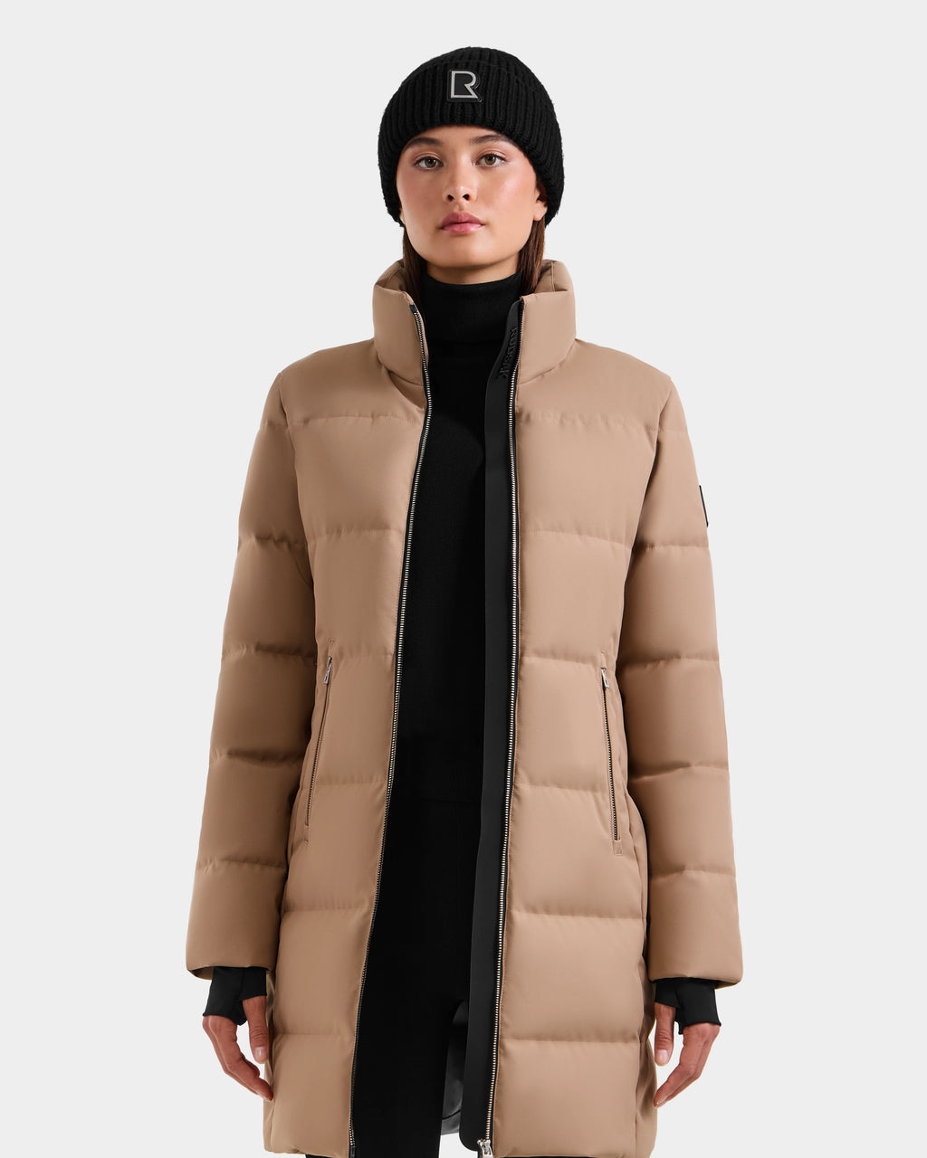 A person faces forward in the Rudsak MIRIAM - L123544 Sand women's down puffer coat with a high collar and front zipper, paired with a black beanie featuring a white R, against a plain light gray background.