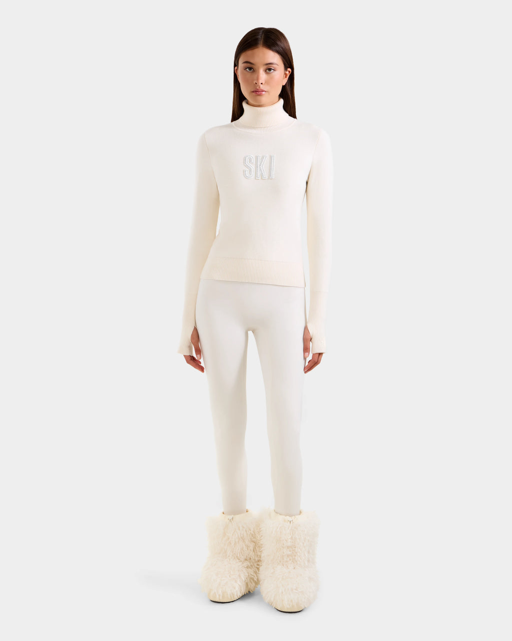 A woman faces forward wearing the Rudsak NOVIE - 8525659 Vanilla all-white après-ski pullover with "SKI" on the front, fitted pants, and large fluffy white boots, set against a plain light gray background.