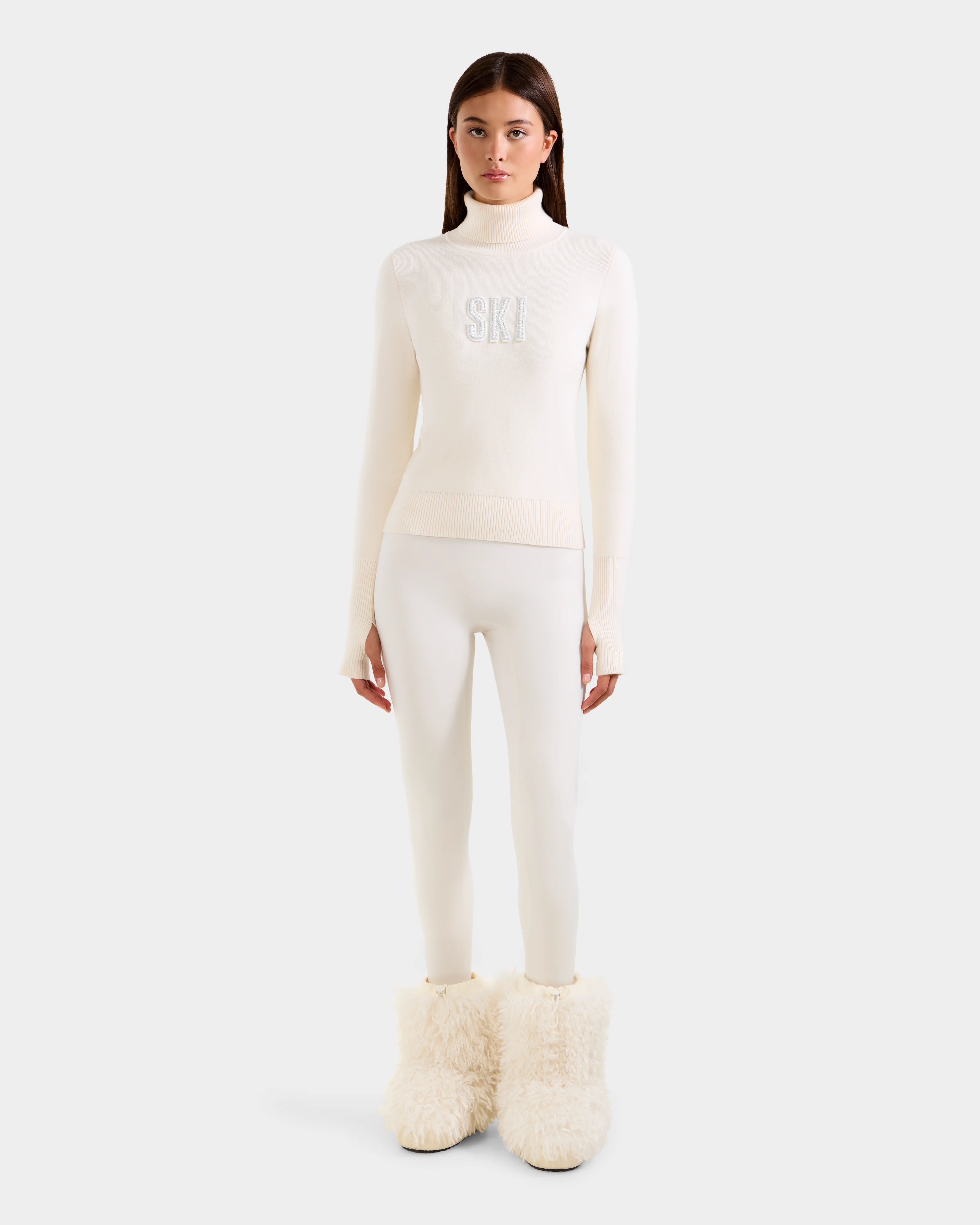 A woman faces forward wearing the Rudsak NOVIE - 8525659 Vanilla all-white après-ski pullover with "SKI" on the front, fitted pants, and large fluffy white boots, set against a plain light gray background.