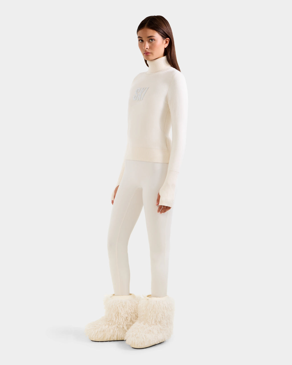 A woman with straight brown hair and a neutral expression wears the Rudsak NOVIE - 8525659 Vanilla, an all-white knit turtleneck sweater, along with slim white pants and oversized fluffy boots, posing against a plain background.