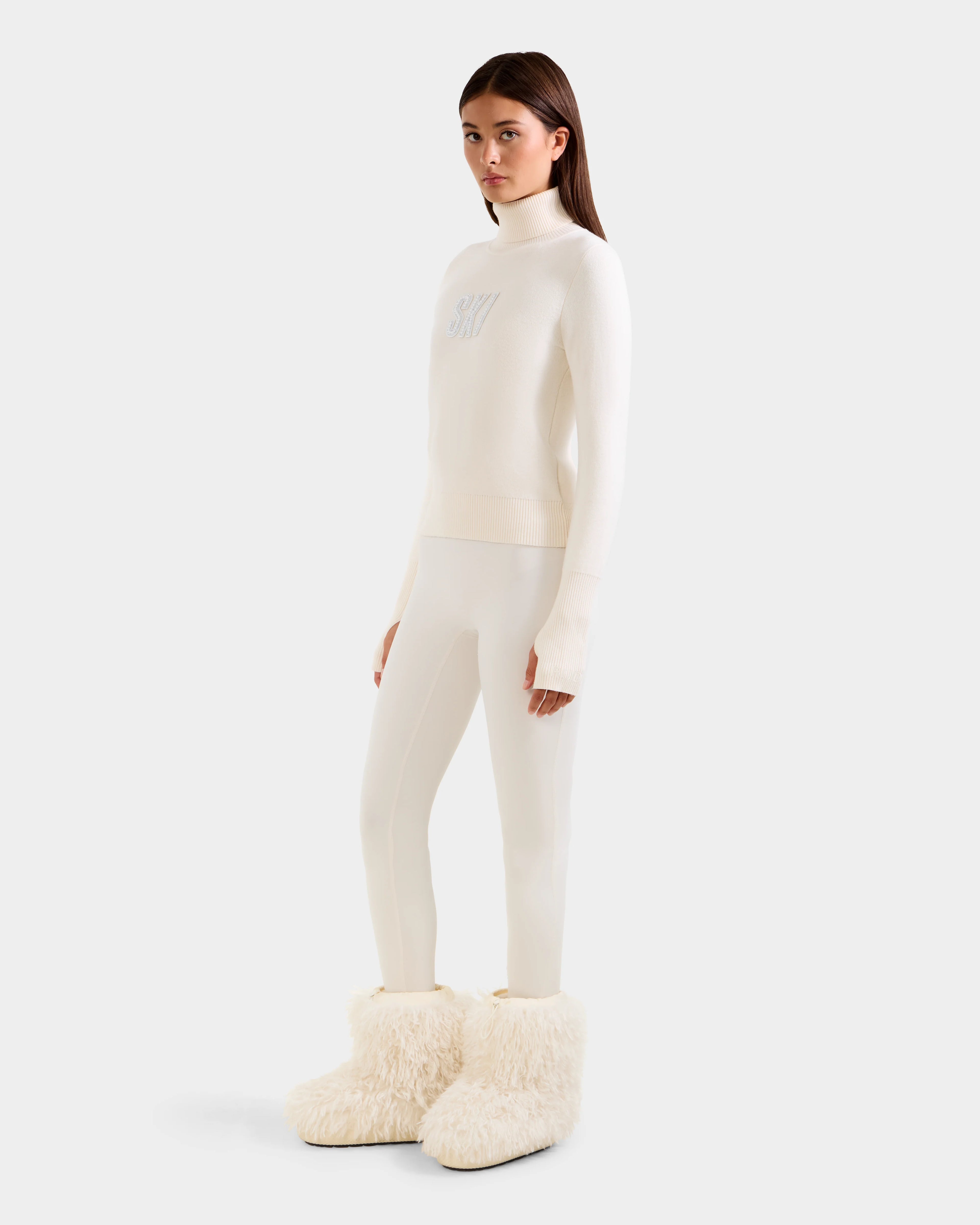 A woman with straight brown hair and a neutral expression wears the Rudsak NOVIE - 8525659 Vanilla, an all-white knit turtleneck sweater, along with slim white pants and oversized fluffy boots, posing against a plain background.