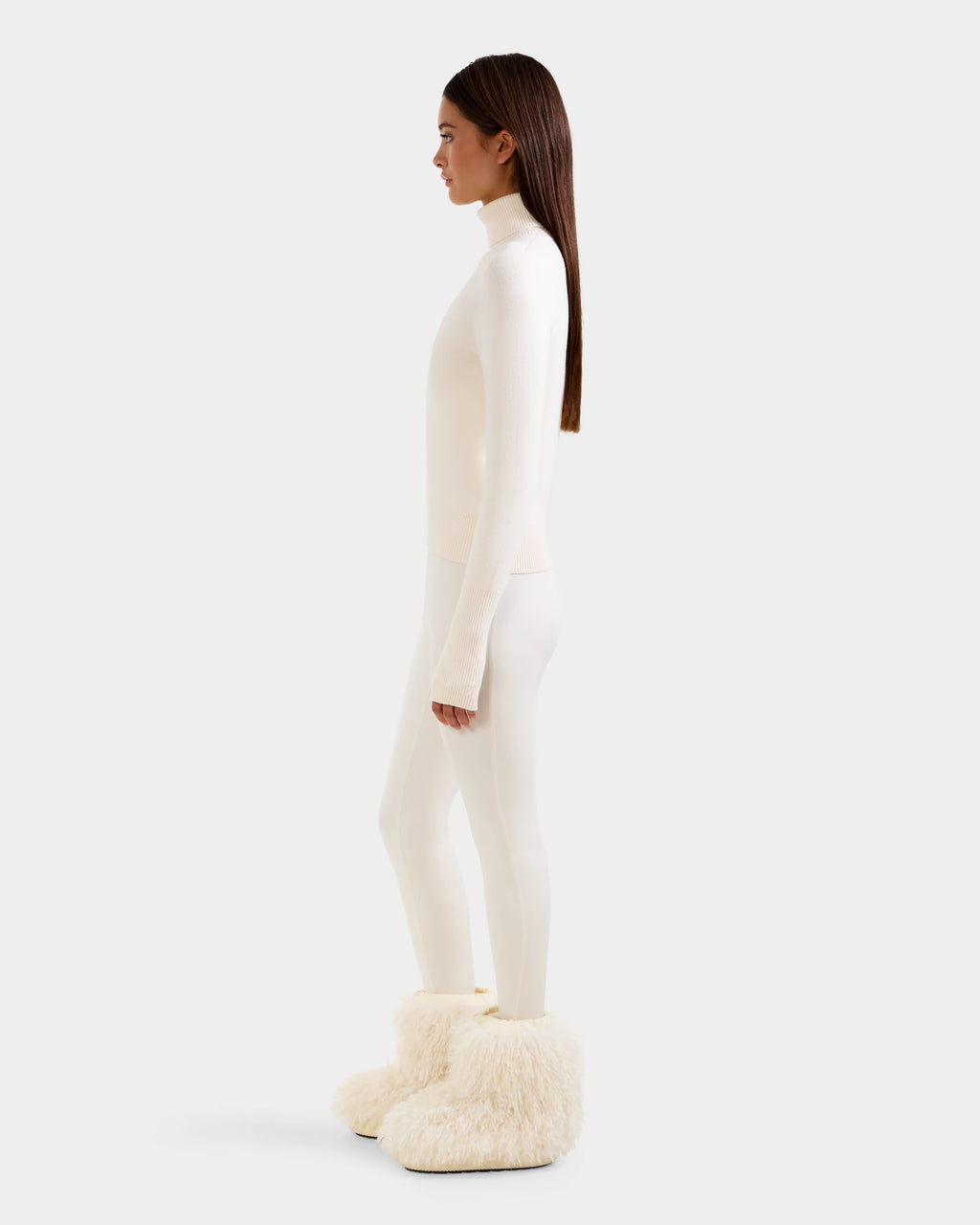 A woman in profile against a white background wears Rudsak's NOVIE - 8525659 Vanilla boots, a sleek women's knit turtleneck, white leggings, and her loose straight brown hair.