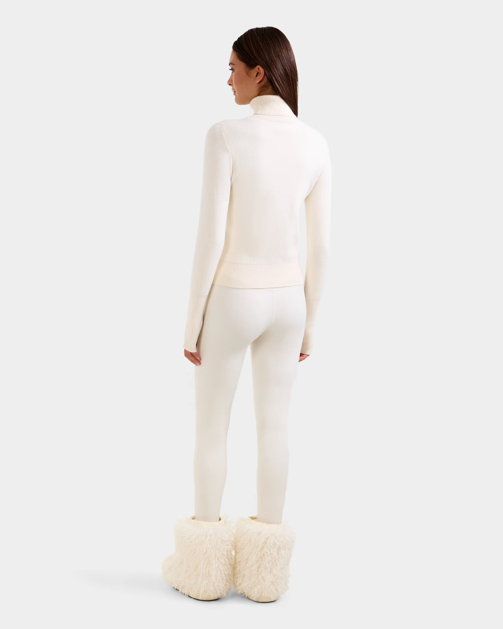 A woman faces away, wearing the Rudsak NOVIE - 8525659 Vanilla women's knit turtleneck sweater with matching leggings and fluffy cream-colored boots, set against a plain light gray background.