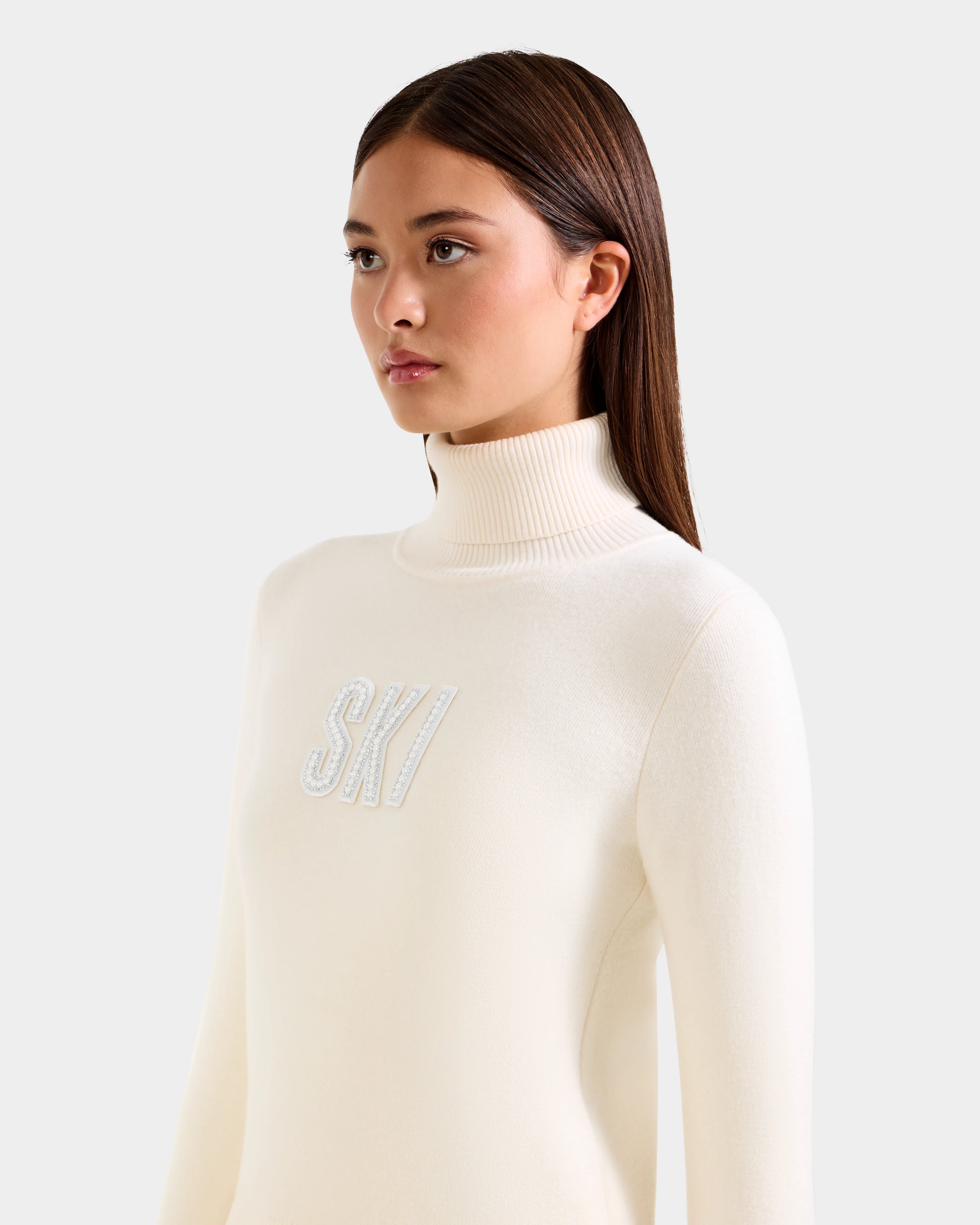 A woman with straight brown hair models the Rudsak NOVIE - 8525659 Vanilla women's knit turtleneck sweater with "SKI" embroidered in white, standing against a light gray background for a chic après-ski look.