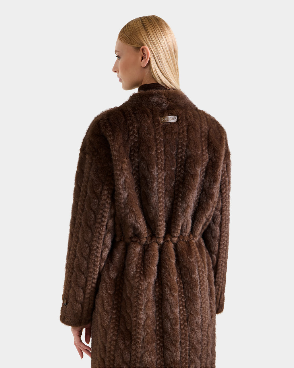 A woman with straight blonde hair wears the Rudsak ORIANA - 8125545 Mocha, a long, dark brown women’s faux fur coat with a textured pattern, belted waist, and a small label on the upper back, seen from behind.