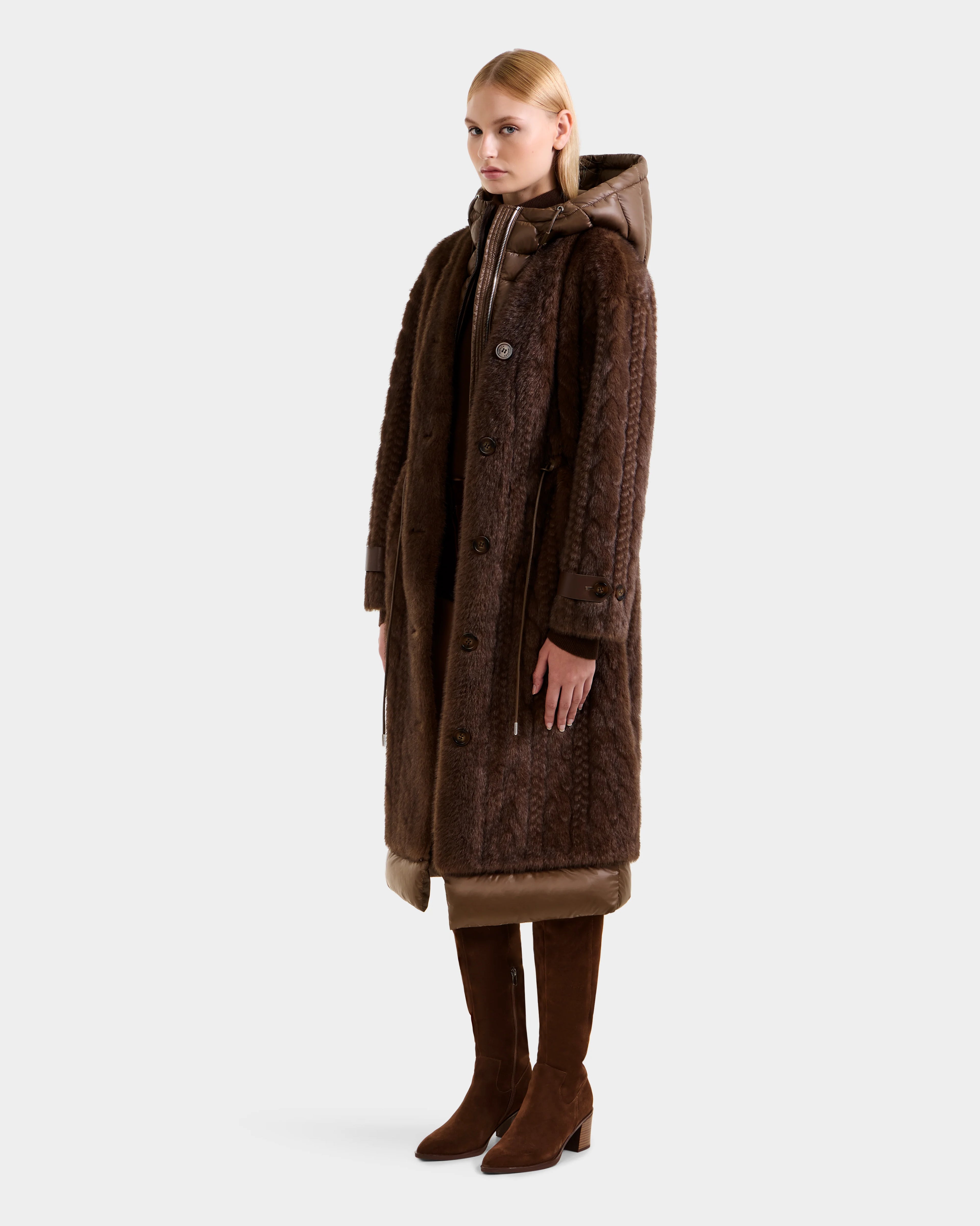 A woman stands facing forward in the ORIANA - 8125545 Mocha coat by Rudsak, featuring vegan leather trims. She pairs it with brown knee-high boots, her arms relaxed at her sides and straight blonde hair, set against a plain light grey background.