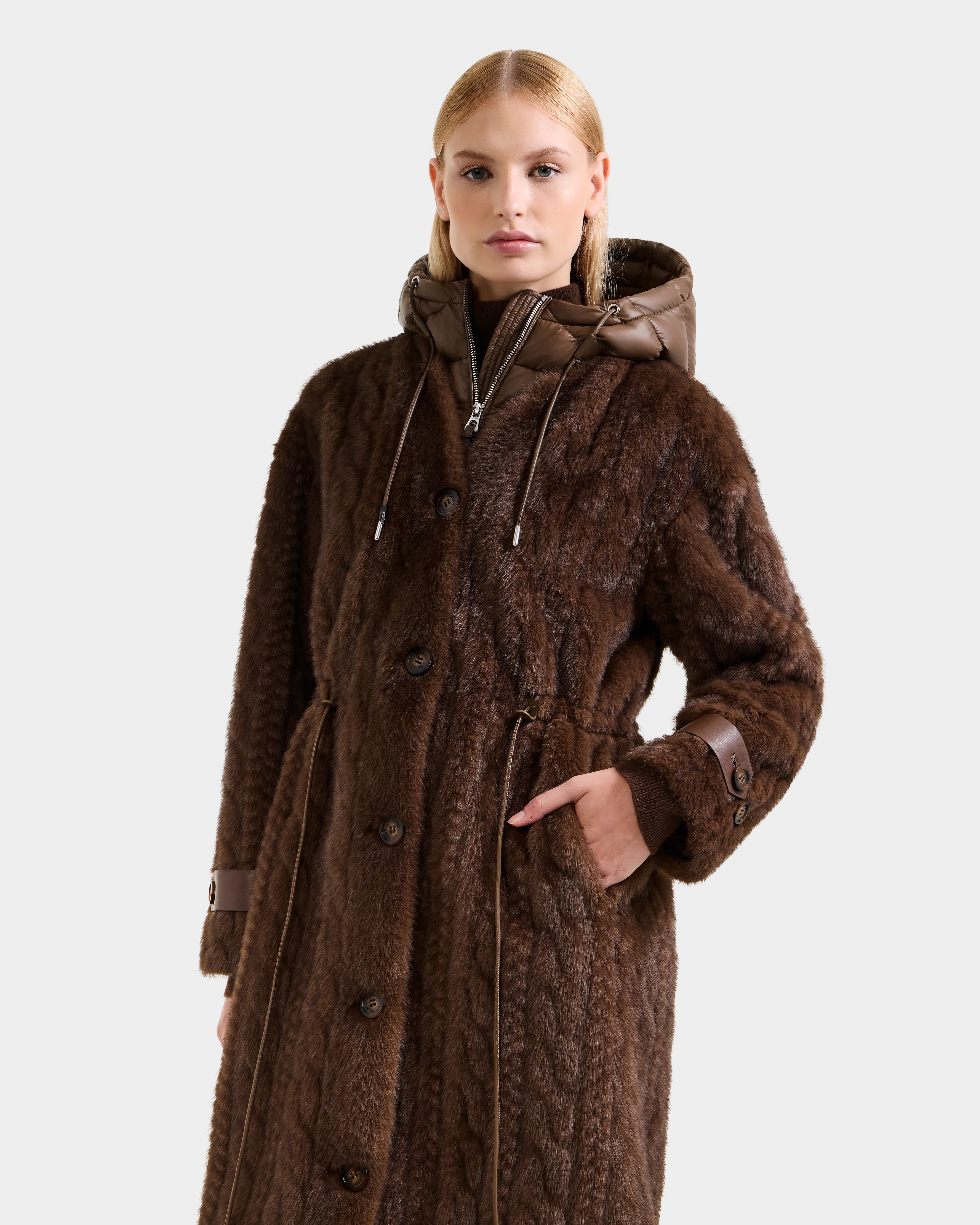 A woman wears the Rudsak ORIANA - 8125545 Mocha, a long dark brown faux fur women’s coat with a hood, button closure, and vegan leather trims, posing with one hand in her pocket against a plain light background.