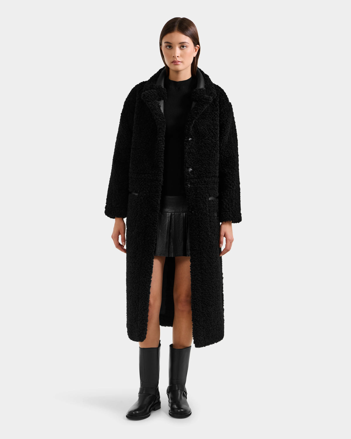 A woman faces forward against a plain light background, dressed in Rudsak's PALOMA - 8125675 Black textured modular long coat over a black top, pleated skirt, and ankle boots.