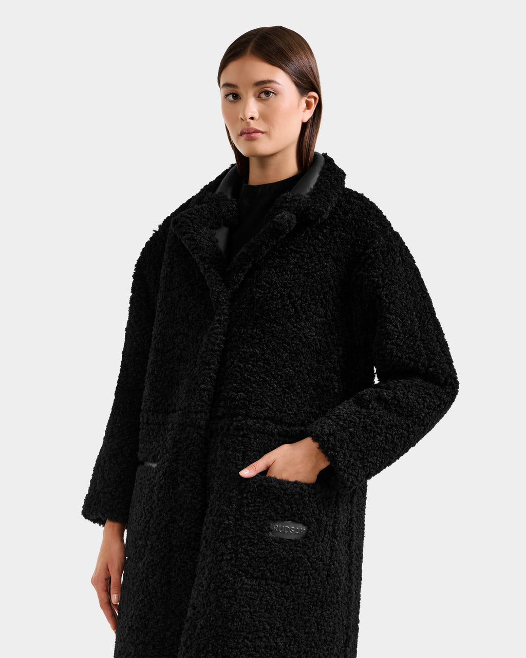 A woman with straight brown hair wears the Rudsak PALOMA - 8125675 Black faux shearling coat with large pockets over a black top, standing against a light gray background.