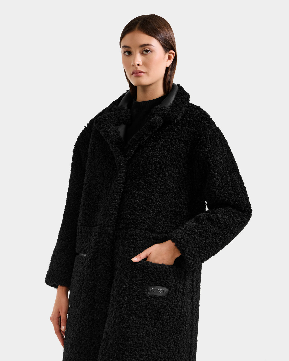 A woman with straight brown hair wears the Rudsak PALOMA - 8125675 Black faux shearling coat with large pockets over a black top, standing against a light gray background.
