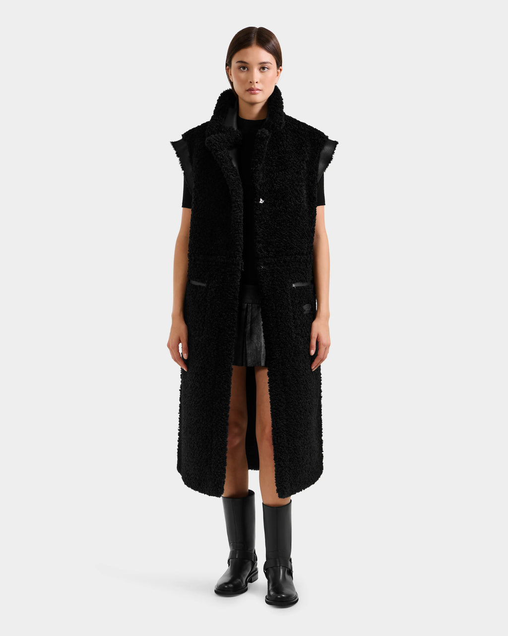 A woman faces forward, wearing the Rudsak PALOMA - 8125675 Black reversible sleeveless coat over a black outfit with knee-high boots, set against a plain light gray background.