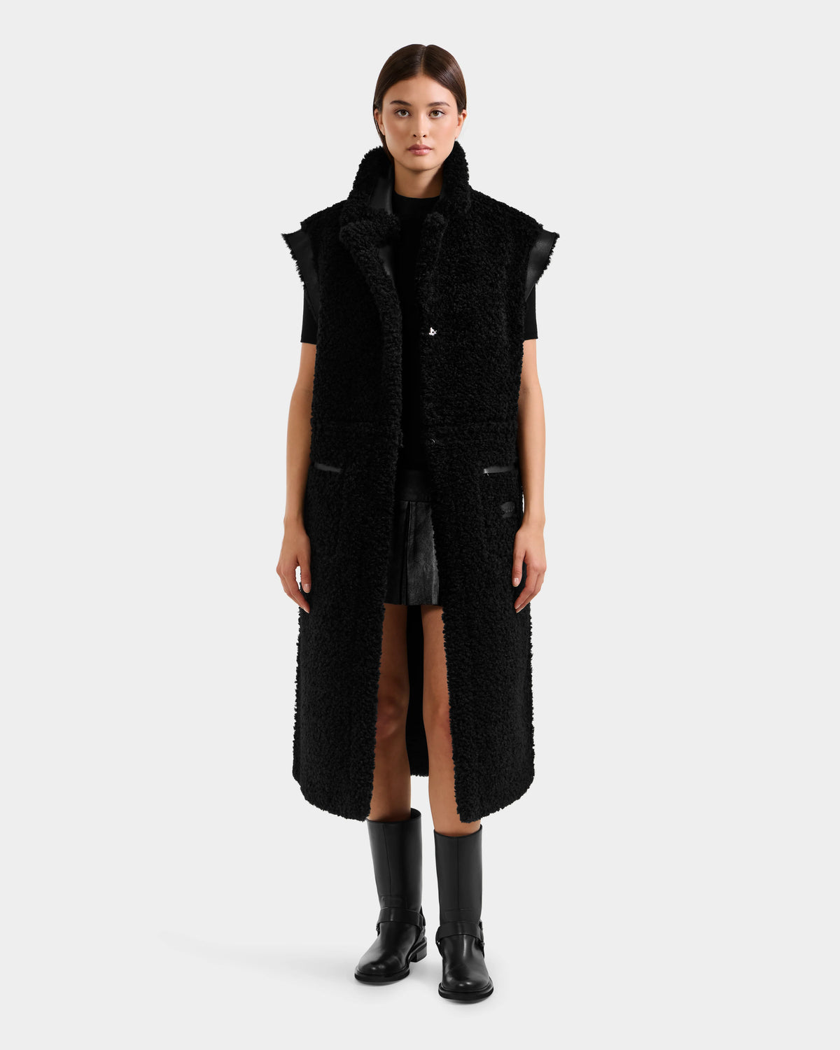 A woman faces forward, wearing the Rudsak PALOMA - 8125675 Black reversible sleeveless coat over a black outfit with knee-high boots, set against a plain light gray background.