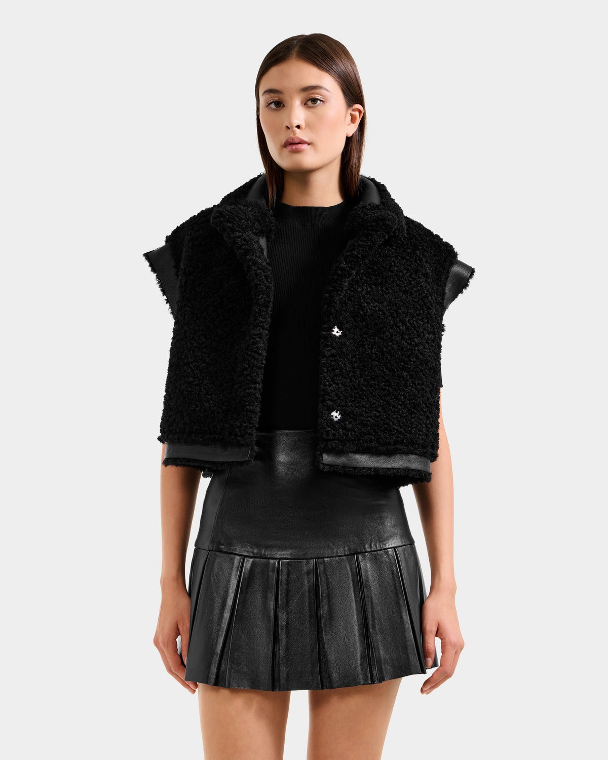 A woman with straight brown hair and a neutral expression wears the Rudsak PALOMA - 8125675 Black textured vest over a black top and pleated leather mini skirt, standing against a plain background.