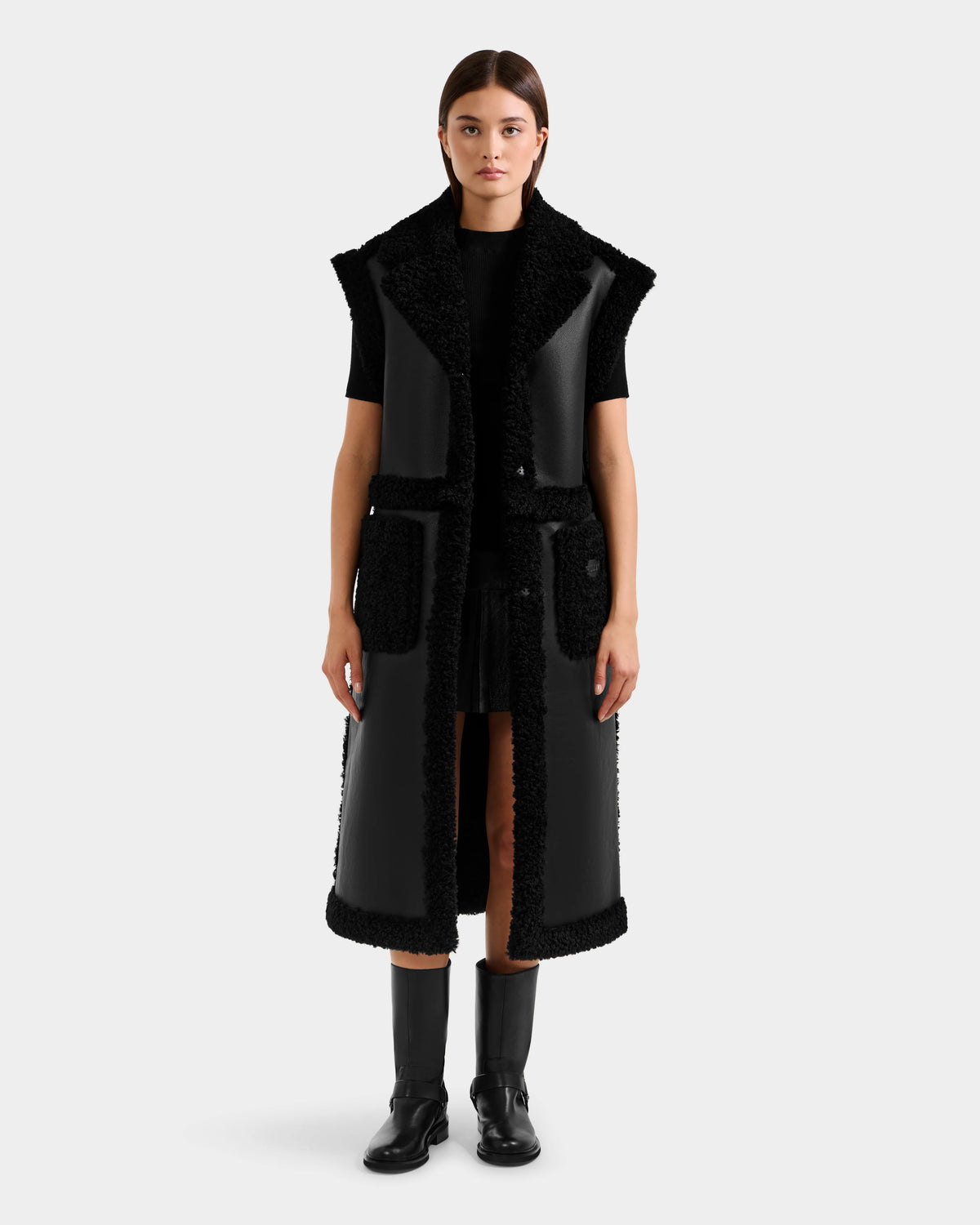 A woman faces forward wearing the Rudsak PALOMA - 8125675 Black faux shearling-trimmed leather coat over a black outfit with knee-high boots, set against a plain light gray background.