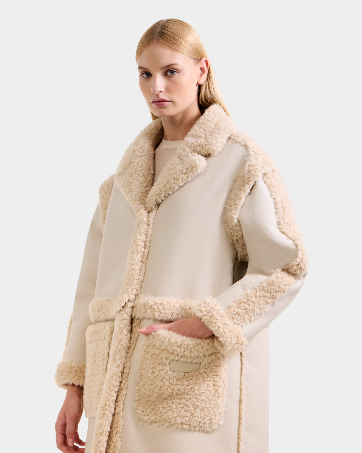 A woman with straight blonde hair models the Rudsak PALOMA - 8125675 Vanilla beige faux shearling coat, featuring fluffy textured trim on the collar, sleeves, front, and pockets, against a plain light gray background.