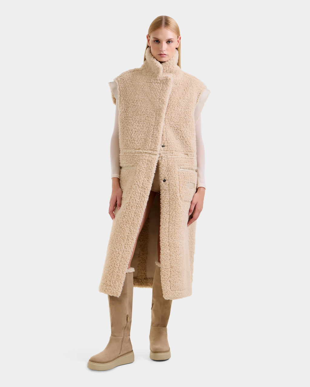 A woman faces forward against a white background, wearing Rudsak's PALOMA - 8125675 Vanilla sleeveless long faux shearling vest over a light top, paired with matching beige knee-high boots. Her hair is straight and sleek.