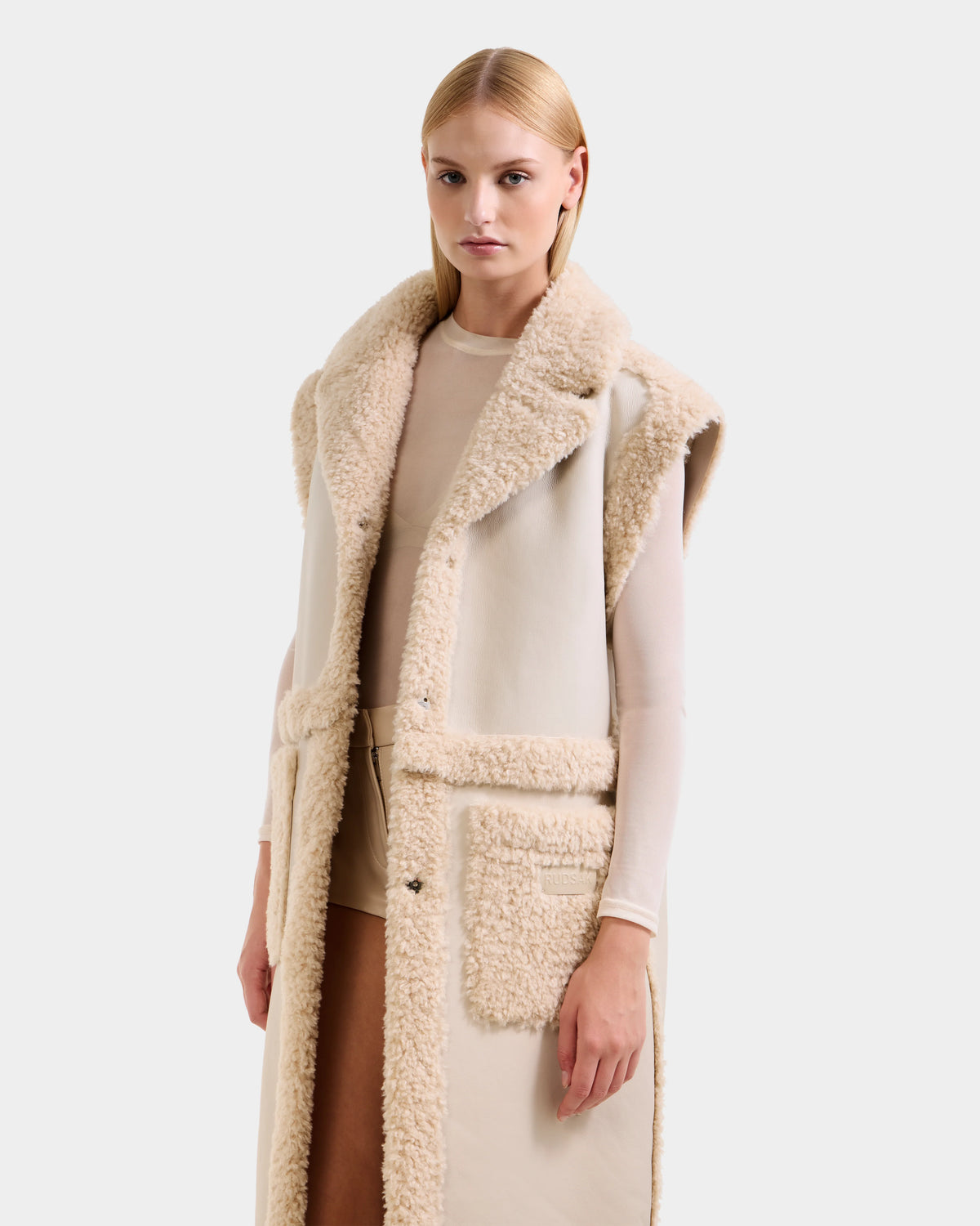 A woman with straight blonde hair models the Rudsak PALOMA - 8125675 Vanilla, a long, sleeveless faux shearling beige coat, layered over a sheer white top and beige shorts against a plain light background.