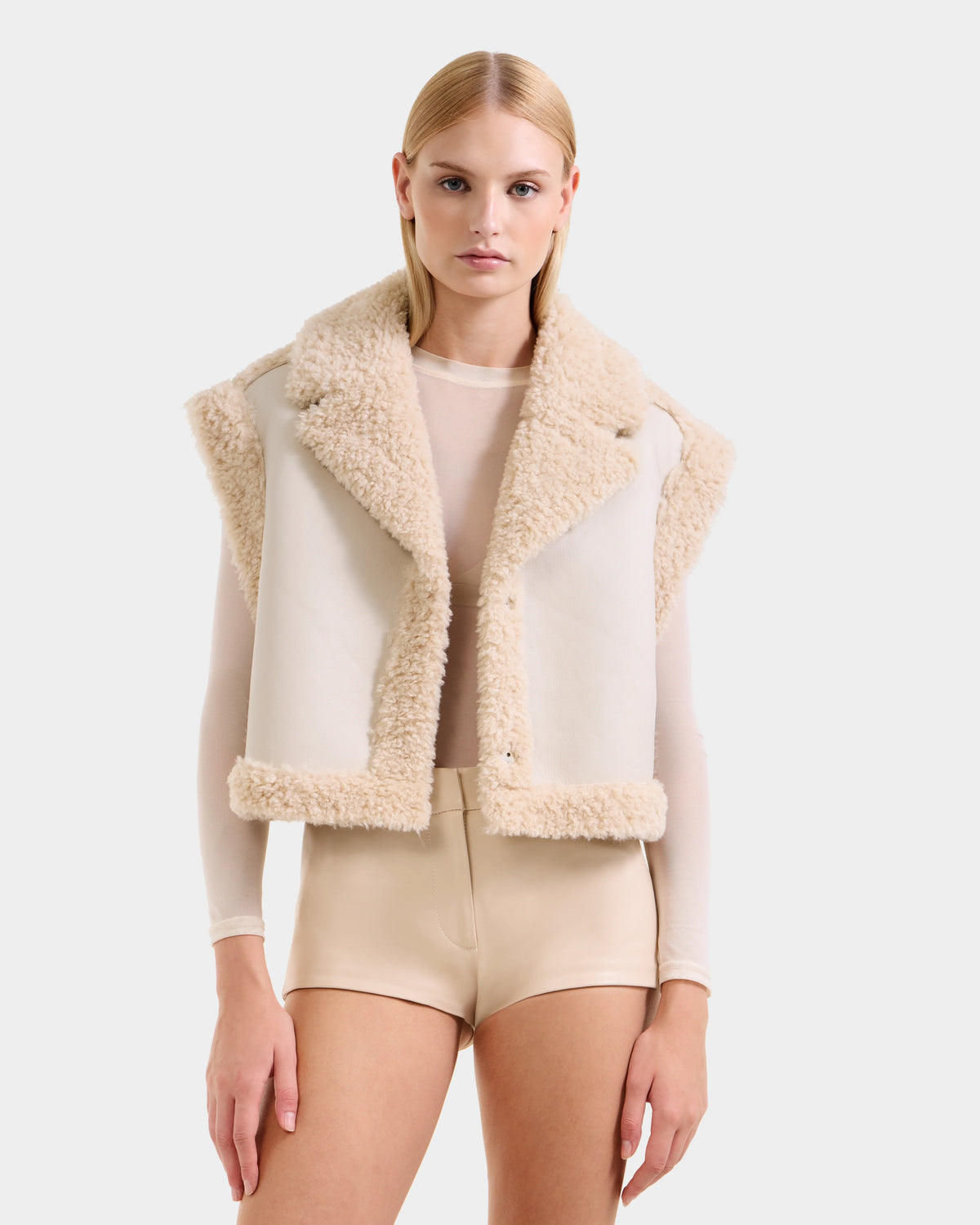 A woman with straight blonde hair and a neutral expression wears the Rudsak PALOMA - 8125675 Vanilla cream faux shearling vest over a sheer long-sleeve top and high-waisted beige shorts, standing against a plain background.