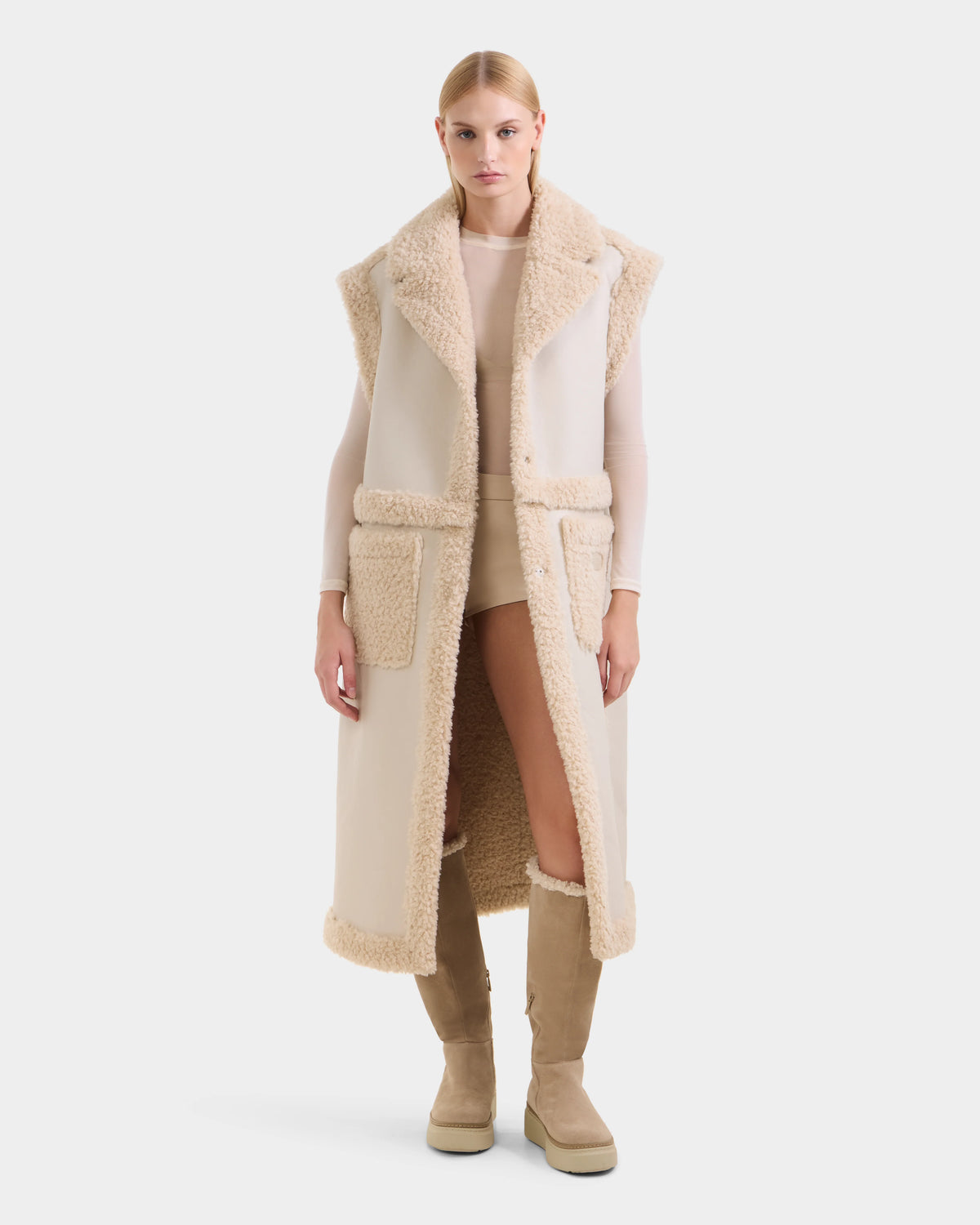 A woman faces forward wearing the Rudsak PALOMA - 8125675 Vanilla faux shearling vest over a beige top and shorts with tan knee-high boots, all against a plain white background that highlights this stylish, modular outerwear piece.