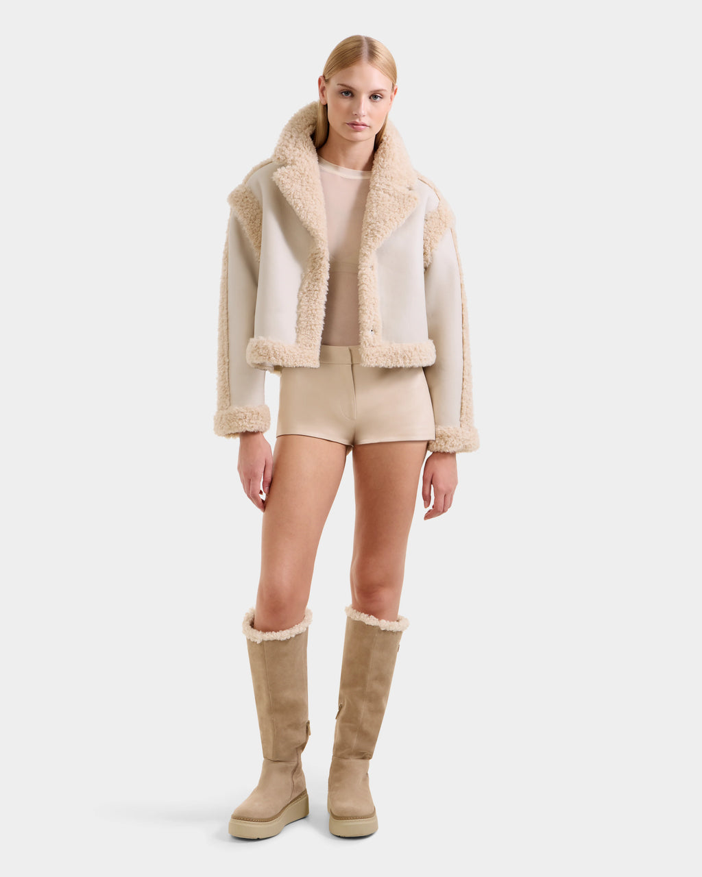 A woman stands against a plain background wearing the Rudsak PALOMA - 8125675 Vanilla jacket, beige shorts, a sheer beige top, and knee-high tan suede boots with shearling trim, looking directly at the camera with a neutral expression.