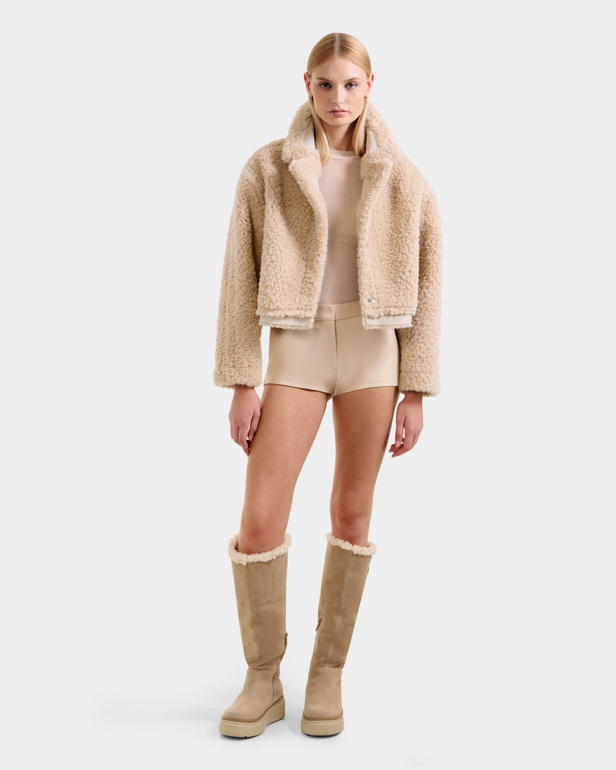 A woman with straight blonde hair and a neutral expression wears the Rudsak PALOMA - 8125675 Vanilla faux shearling modular jacket, nude top, matching high-waisted shorts, and tall tan shearling boots against a plain white background.
