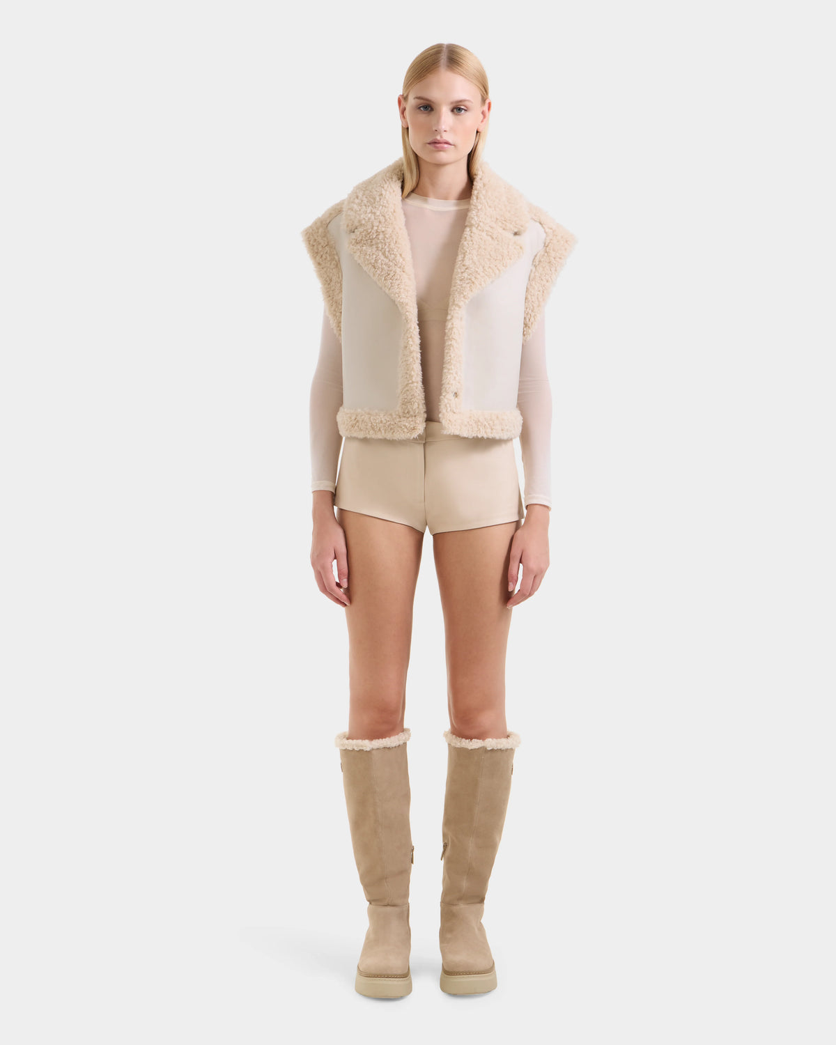 A woman faces forward against a white background wearing the Rudsak PALOMA - 8125675 Vanilla beige faux shearling vest, a sheer long-sleeve top, beige shorts, and knee-high tan boots with shearling trim.