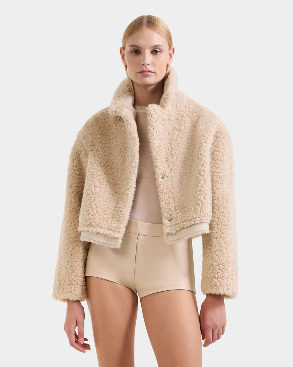 A person wears the Rudsak PALOMA - 8125675 Vanilla faux shearling cropped jacket over a sheer beige top and high-waisted beige shorts, standing facing forward against a plain white background.