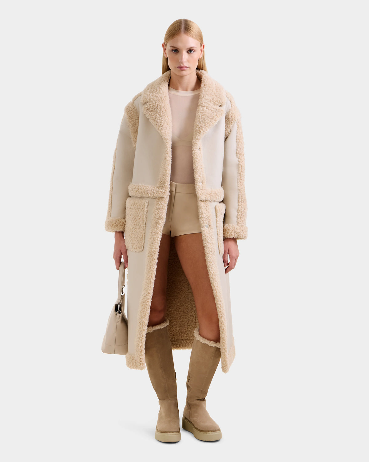 A woman faces forward in the Rudsak PALOMA - 8125675 Vanilla, a long beige faux shearling coat, paired with a sheer top, shorts, knee-high boots, and a beige handbag. The modular outerwear stands out against the plain white background.