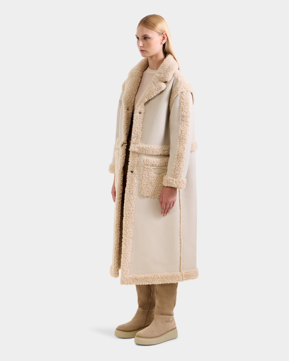 A woman models the Rudsak PALOMA - 8125675 Vanilla faux shearling coat with large lapels and patch pockets, paired with beige suede boots. She stands slightly turned against a plain light background.