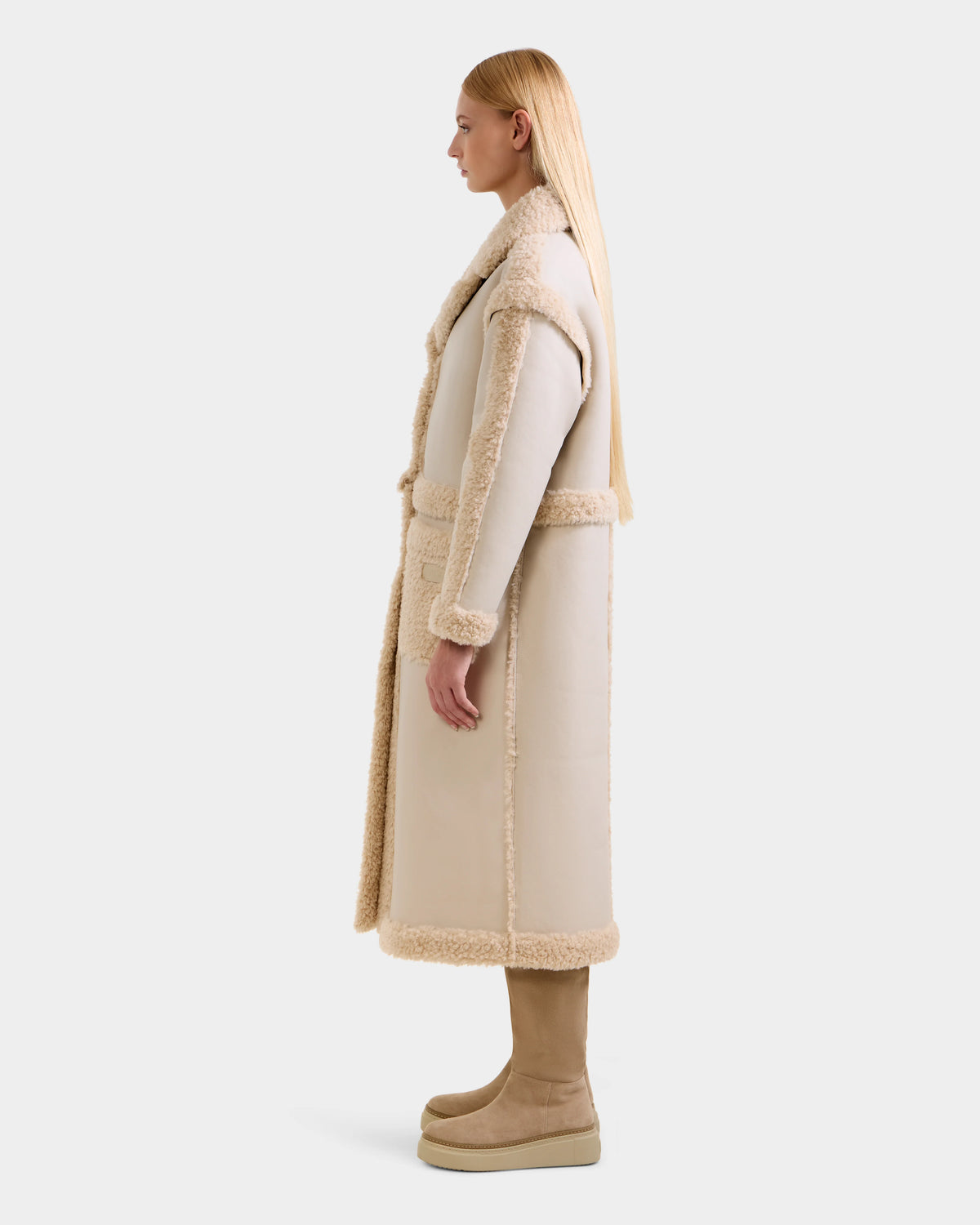 A woman with long straight blonde hair stands in profile, wearing the Rudsak PALOMA - 8125675 Vanilla faux shearling coat with matching beige pants and platform boots against a plain light gray background.