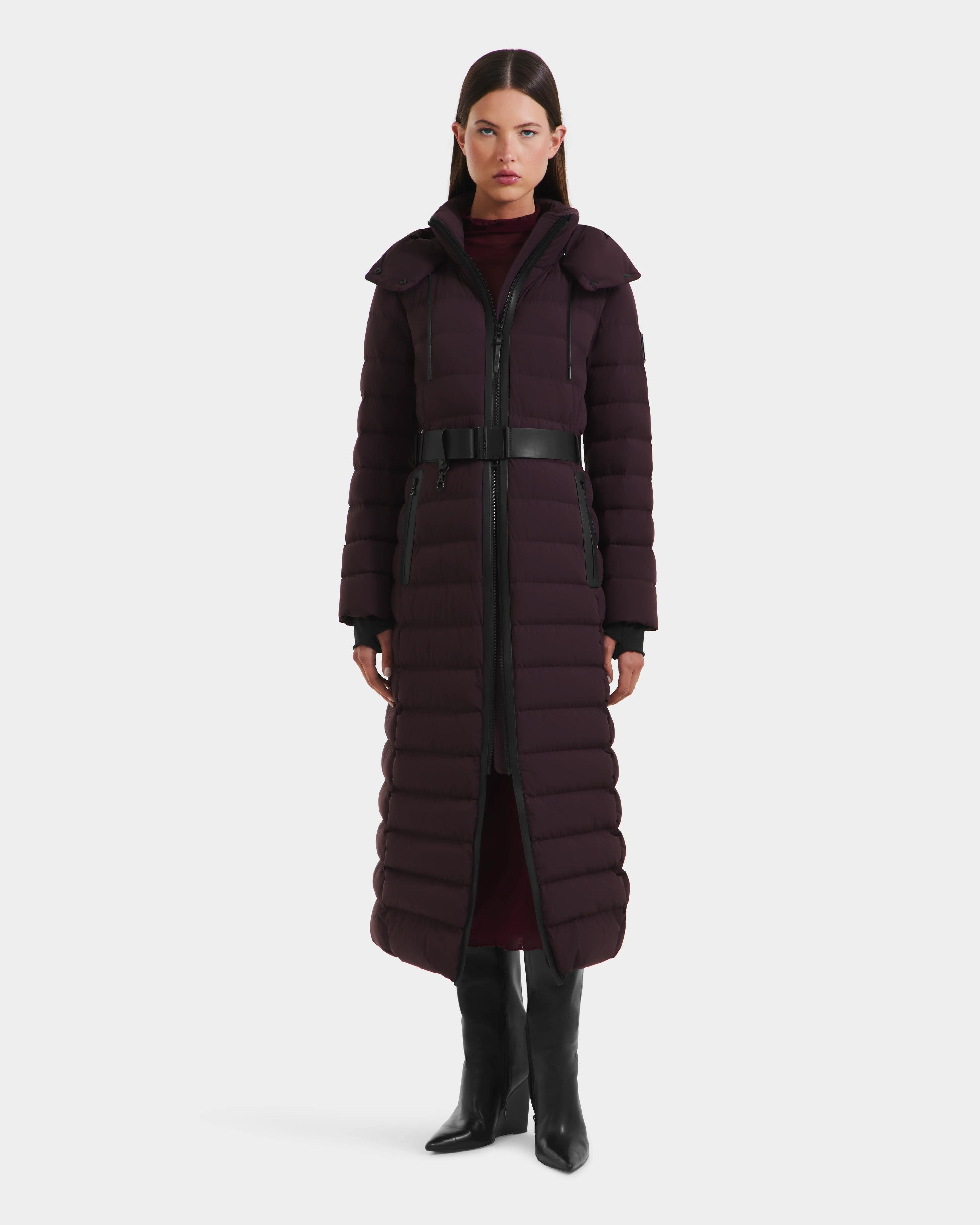 A woman with straight dark hair and a neutral expression wears the Rudsak PELIN L - 8124815 Bordeaux Mocha womens long down puffer, styled with a black belt, knee-high boots, and a high-neck burgundy top.