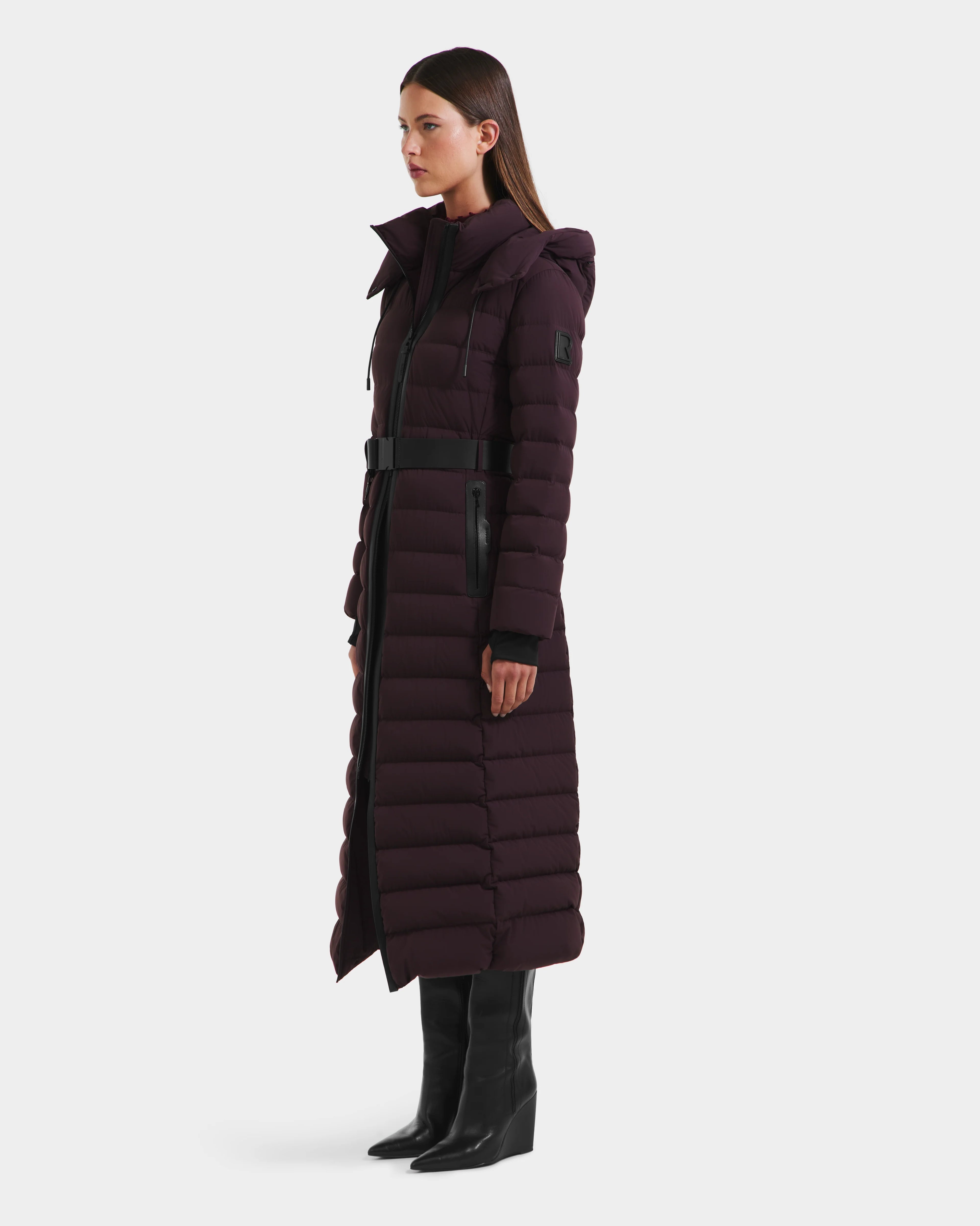 A woman stands in profile wearing the Rudsak PELIN L - 8124815 Bordeaux Mocha, a long down puffer with belted waist and hood, paired with black knee-high boots against a plain white background.