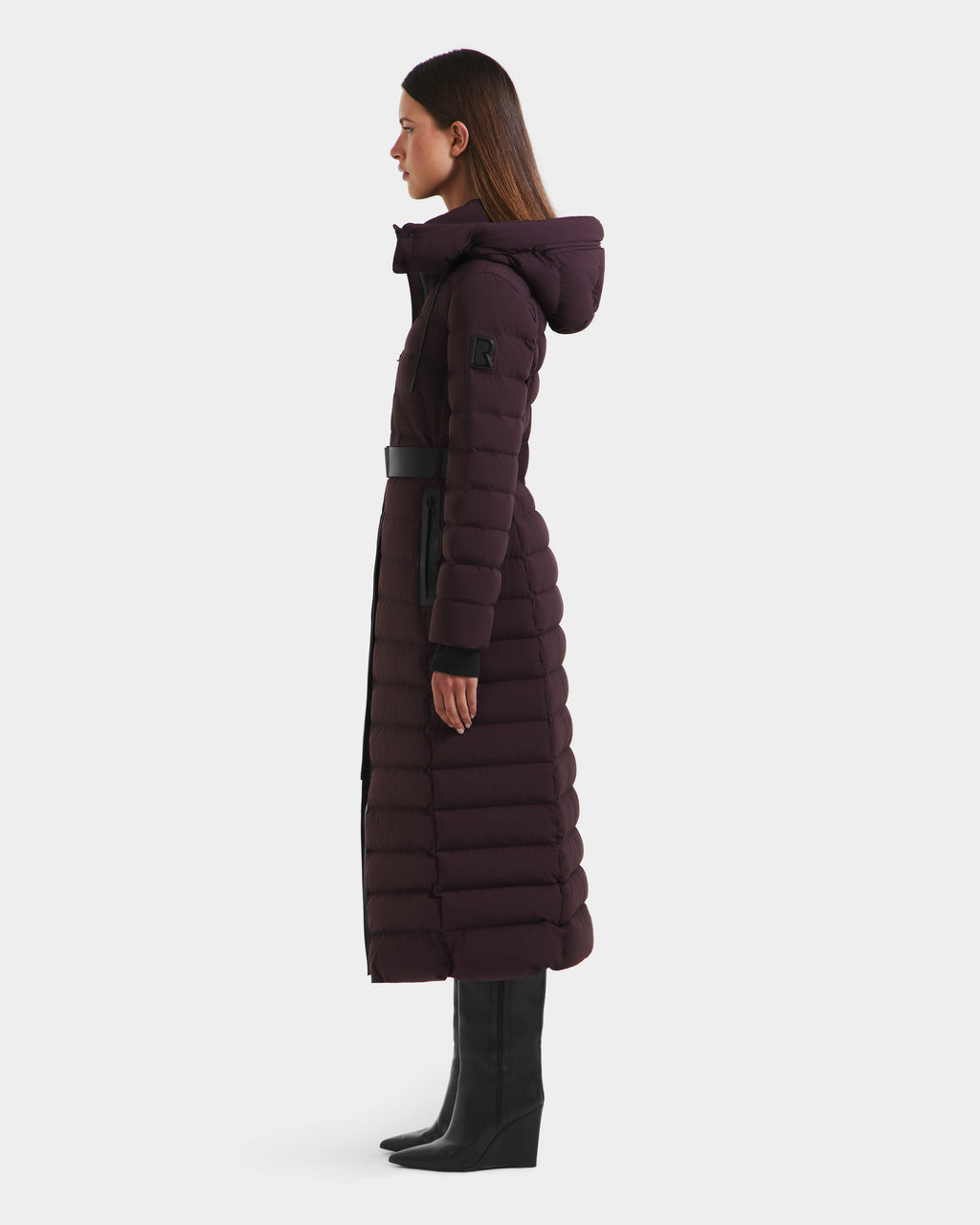 A woman stands in profile wearing the Rudsak PELIN L - 8124815 Bordeaux Mocha long down puffer with a hood, black belt, and black knee-high boots against a plain white background.