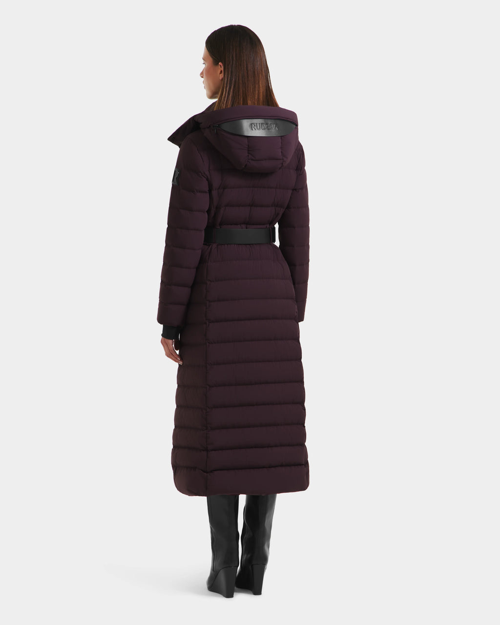 A woman with straight brown hair wears the Rudsak PELIN L - 8124815 Bordeaux Mocha long down puffer with a hood and belt, standing and facing away to show the back of the quilted coat paired with tall black boots.