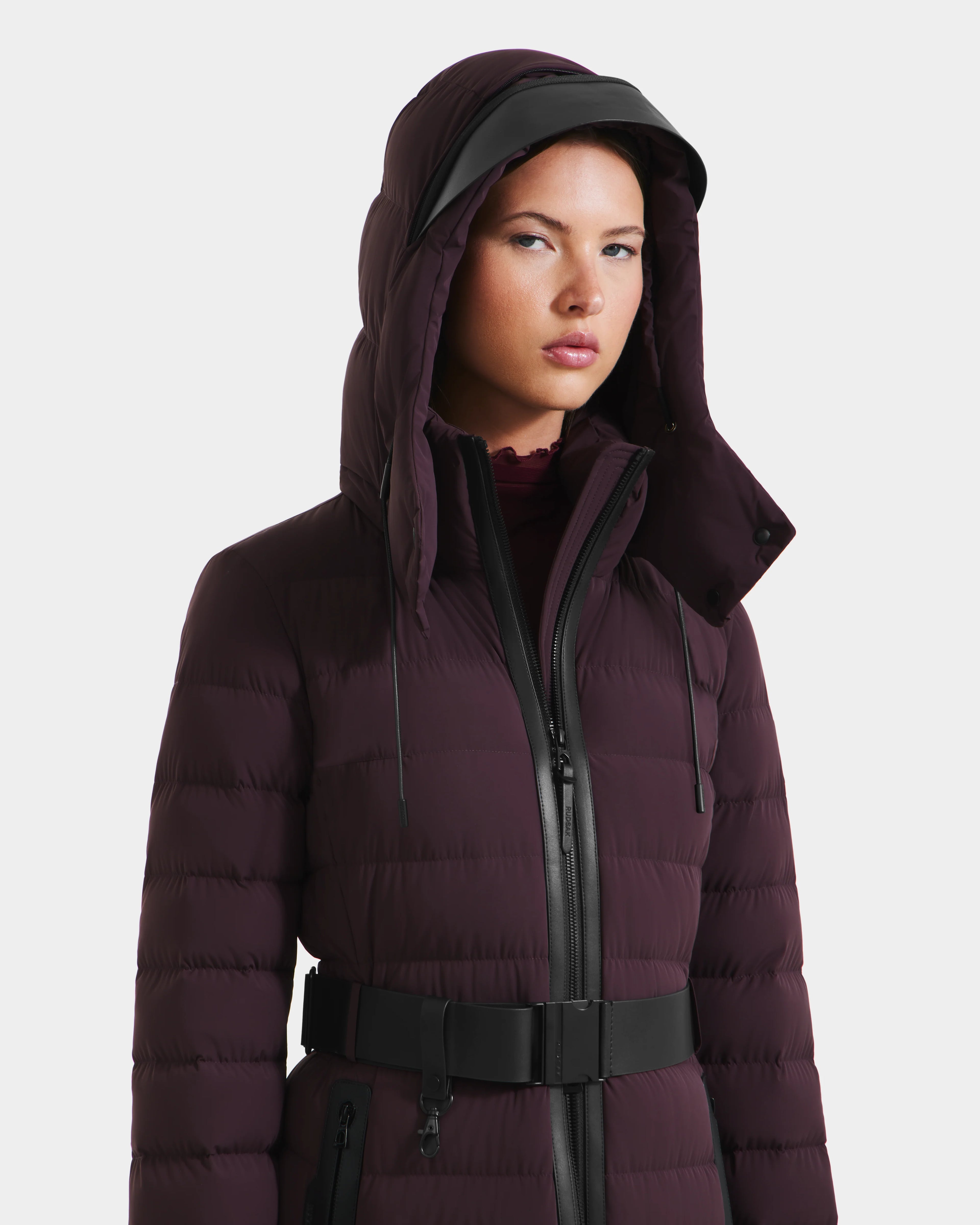 A person wears the Rudsak PELIN L - 8124815 Bordeaux, a dark maroon hooded womens long down puffer with a black zipper and belt, standing against a plain white background.