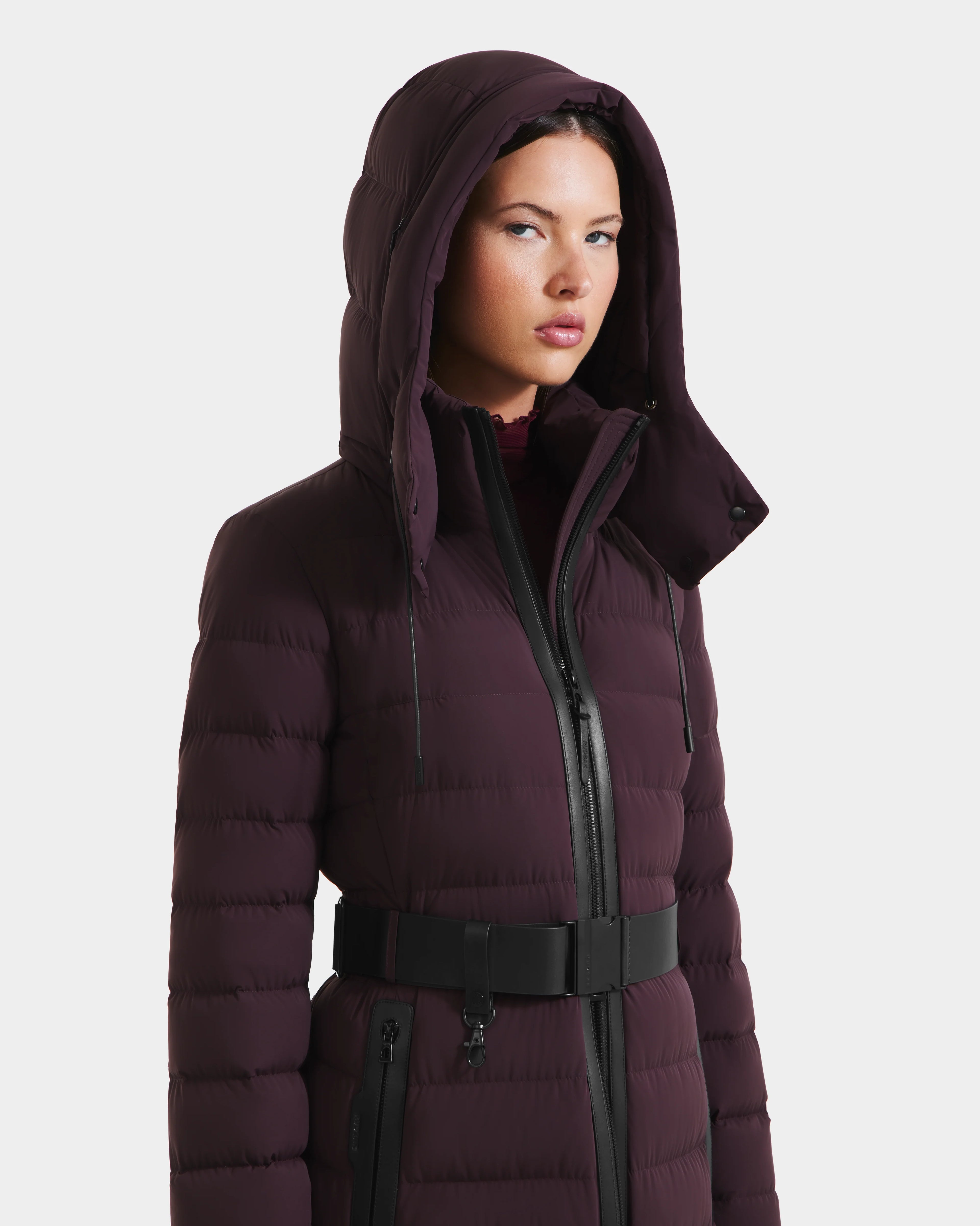 A woman models the Rudsak PELIN L - 8124815 Bordeaux Mocha, a dark purple quilted puffer coat with a hood, black belt, and zipper, standing against a plain white background.