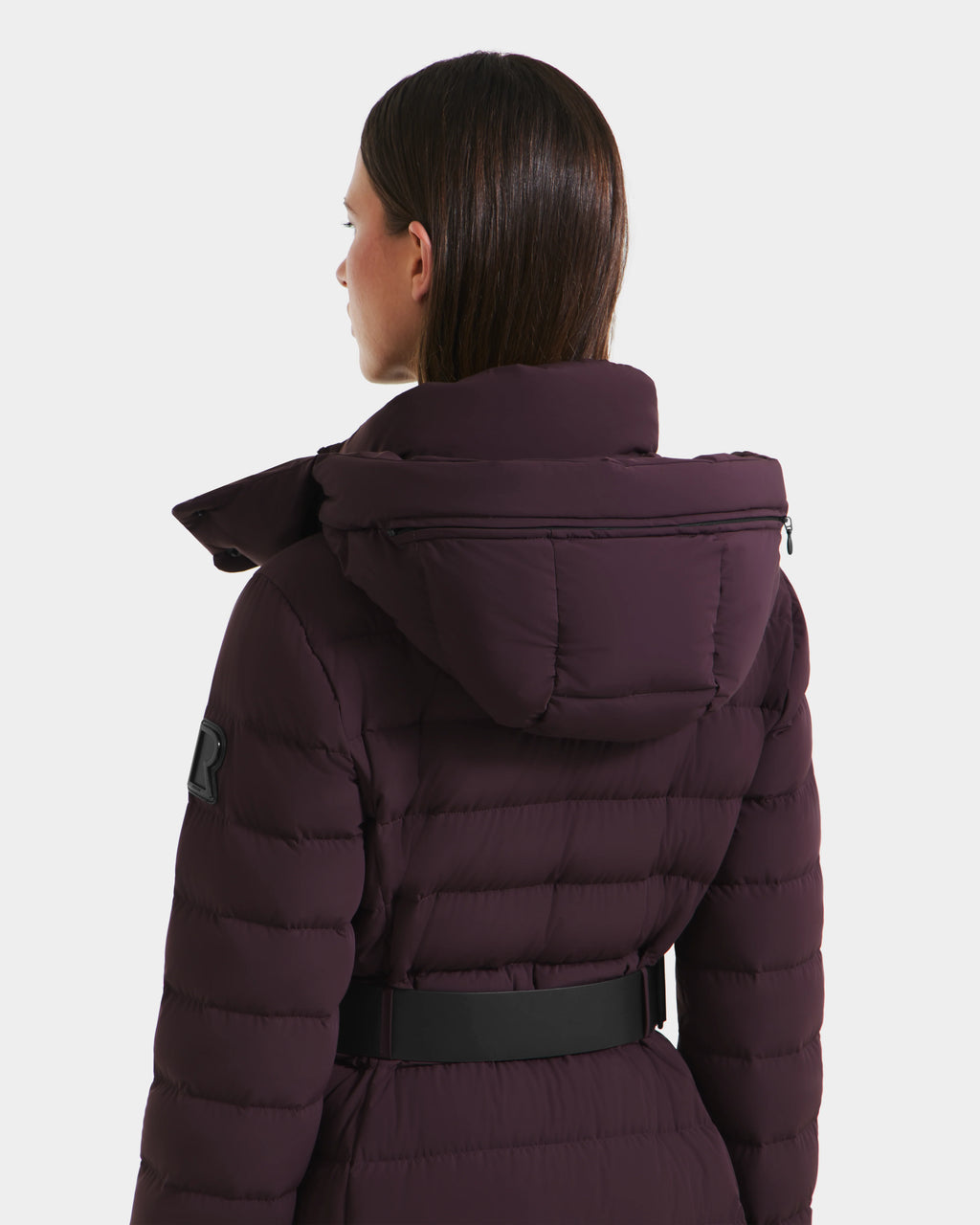 A person with straight brown hair is seen from the back wearing the Rudsak PELIN L - 8124815 Bordeaux Mocha women’s long down puffer jacket with a black belt, against a plain white background.