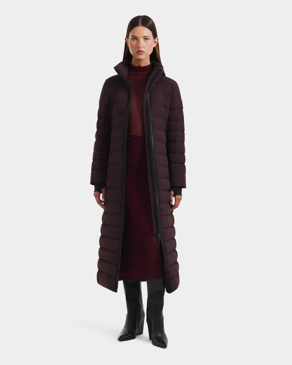 A woman faces forward against a plain white background, wearing the Rudsak PELIN L - 8124815 Bordeaux Mocha long down puffer over a burgundy turtleneck dress and black knee-high boots.