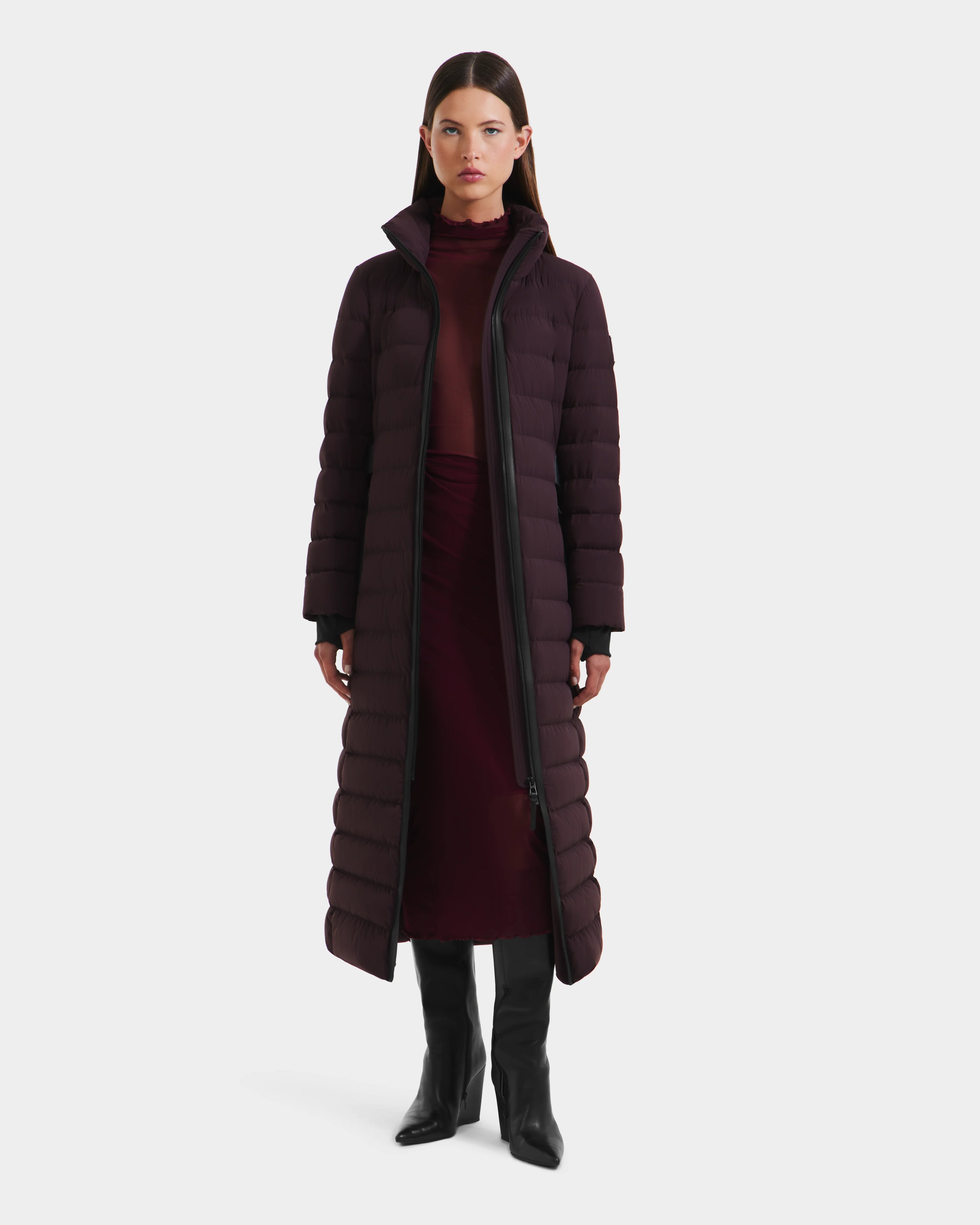A woman faces forward against a plain white background, wearing the Rudsak PELIN L - 8124815 Bordeaux Mocha long down puffer over a burgundy turtleneck dress and black knee-high boots.
