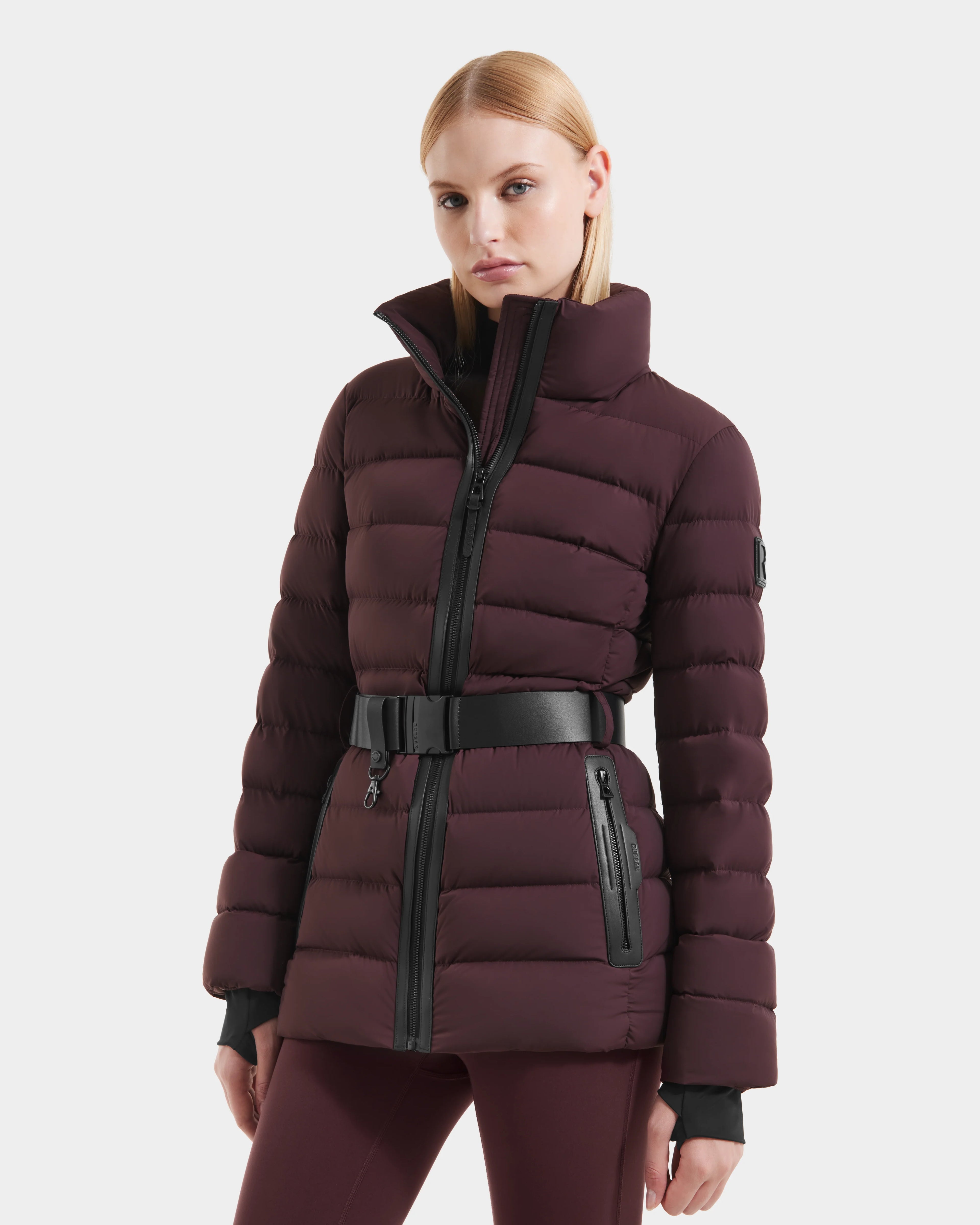 A woman stands against a white background wearing the Rudsak PELIN S - 8124813 Bordeaux down puffer with a high collar, black zipper, and matching belt, her hands at her sides.