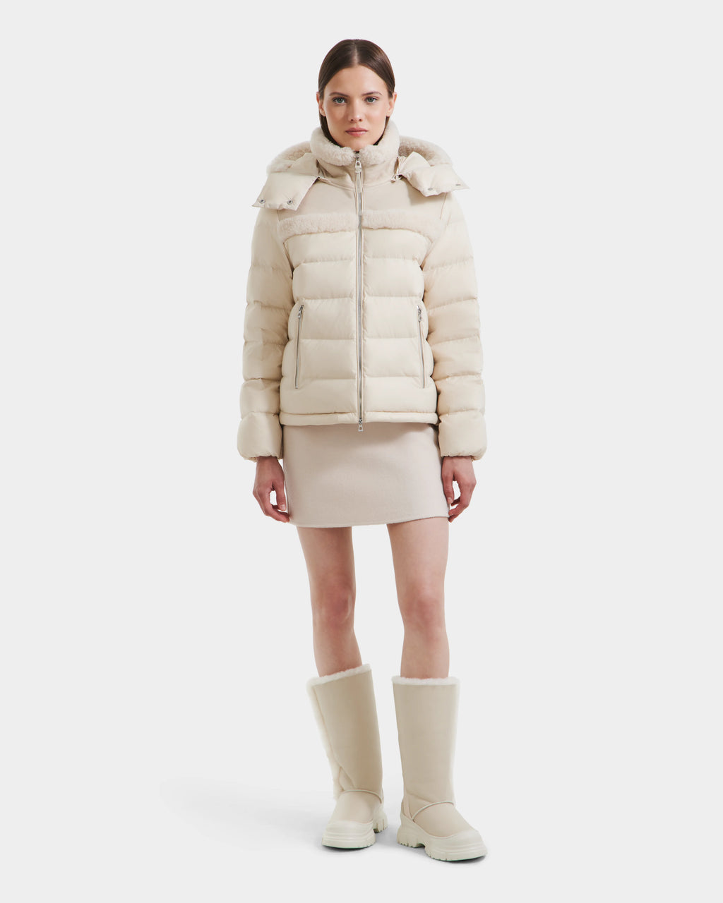 A woman faces forward against a white background wearing the Rudsak SANDRA - 8125612 Vanilla puffer jacket, a matching short skirt, and tall cream boots.