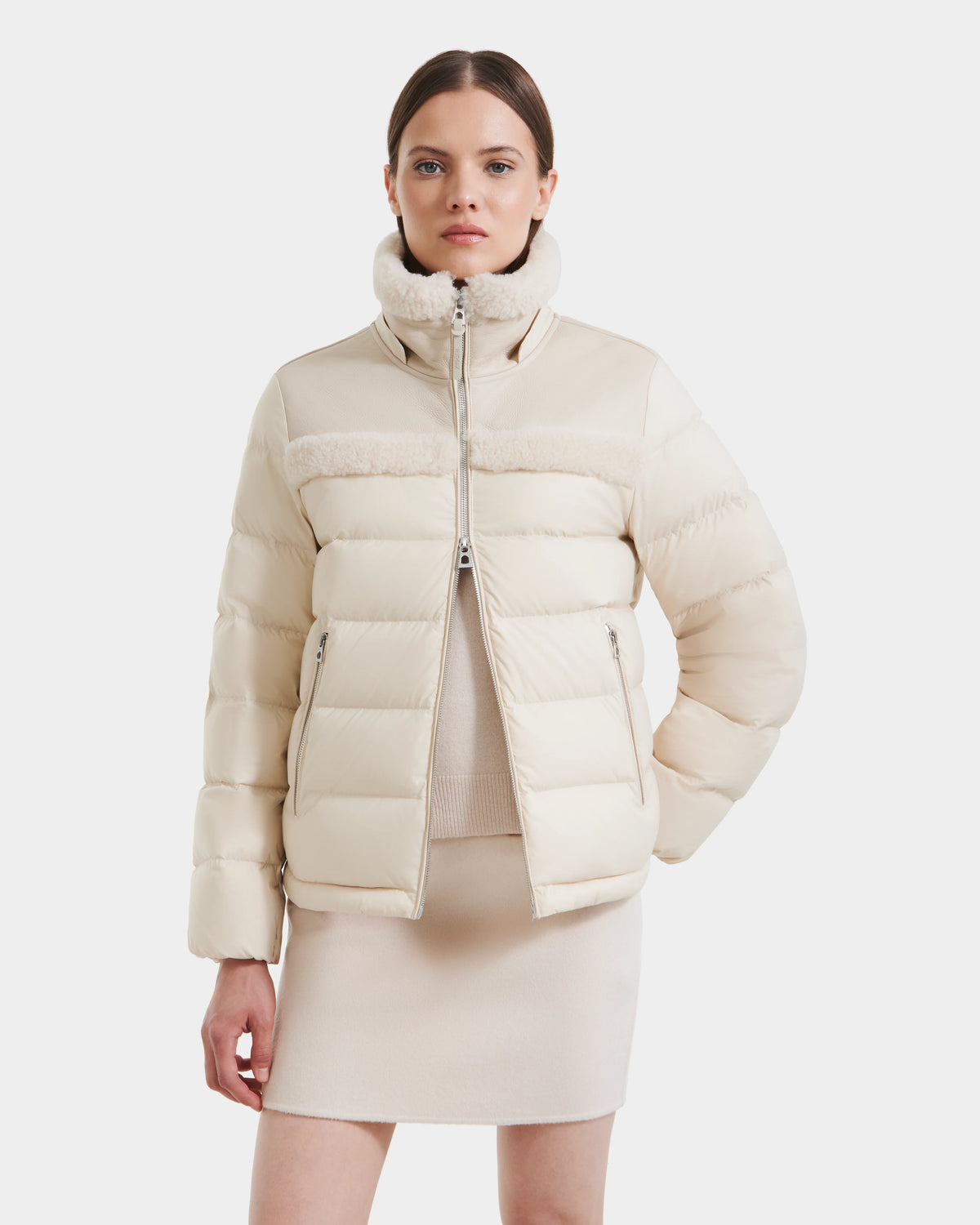 A woman with straight brown hair stands with one hand in her pocket, wearing the Rudsak SANDRA - 8125612 Vanilla shearling puffer jacket. She styles its high collar with a matching skirt and top for a modern, coordinated look.
