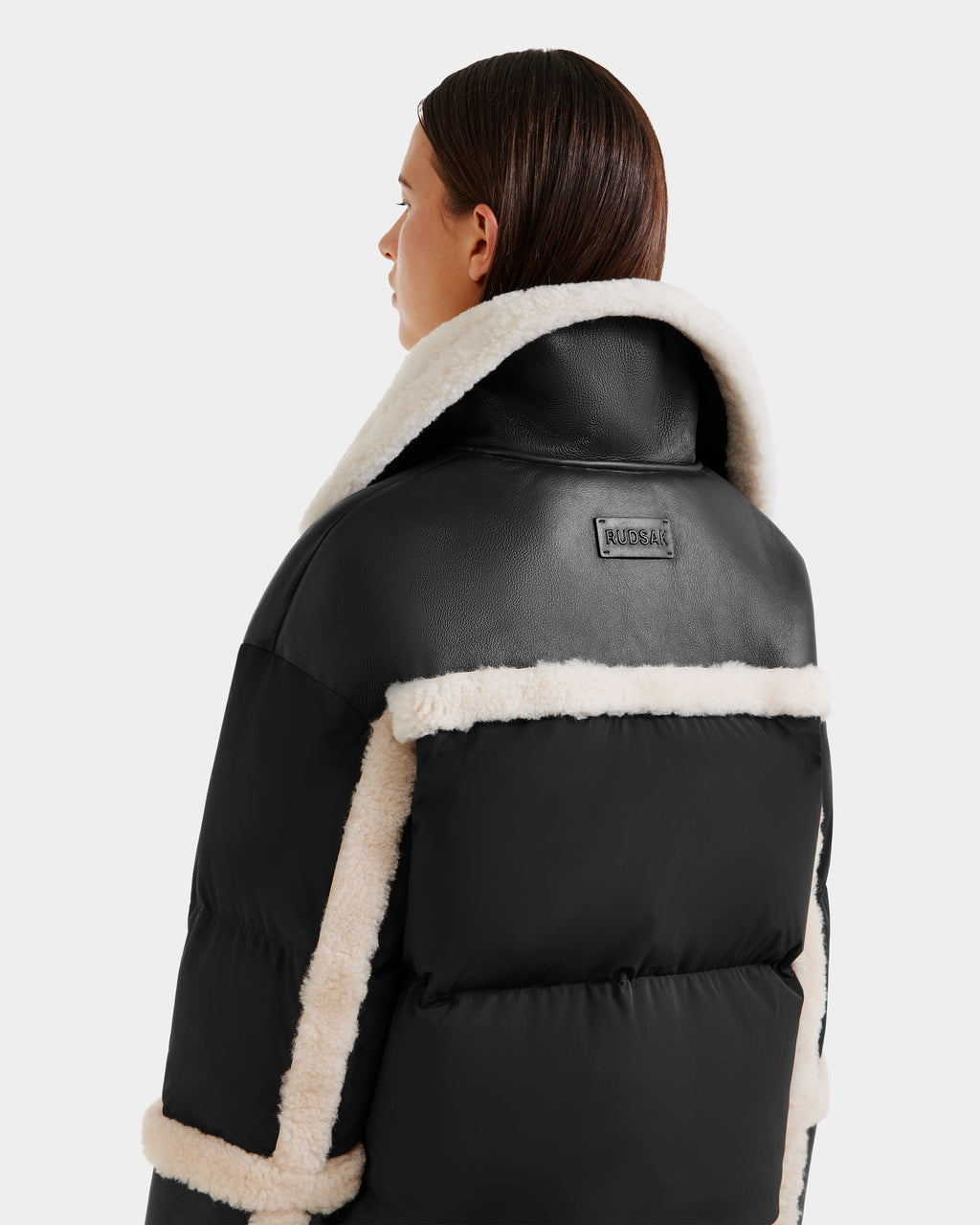 A woman with straight brown hair models the Rudsak SHIRLEY - 8125613 Black jacket, featuring white faux shearling trim on the collar, back, and sleeves plus a branded rectangular patch on the back. She is facing away from the camera.