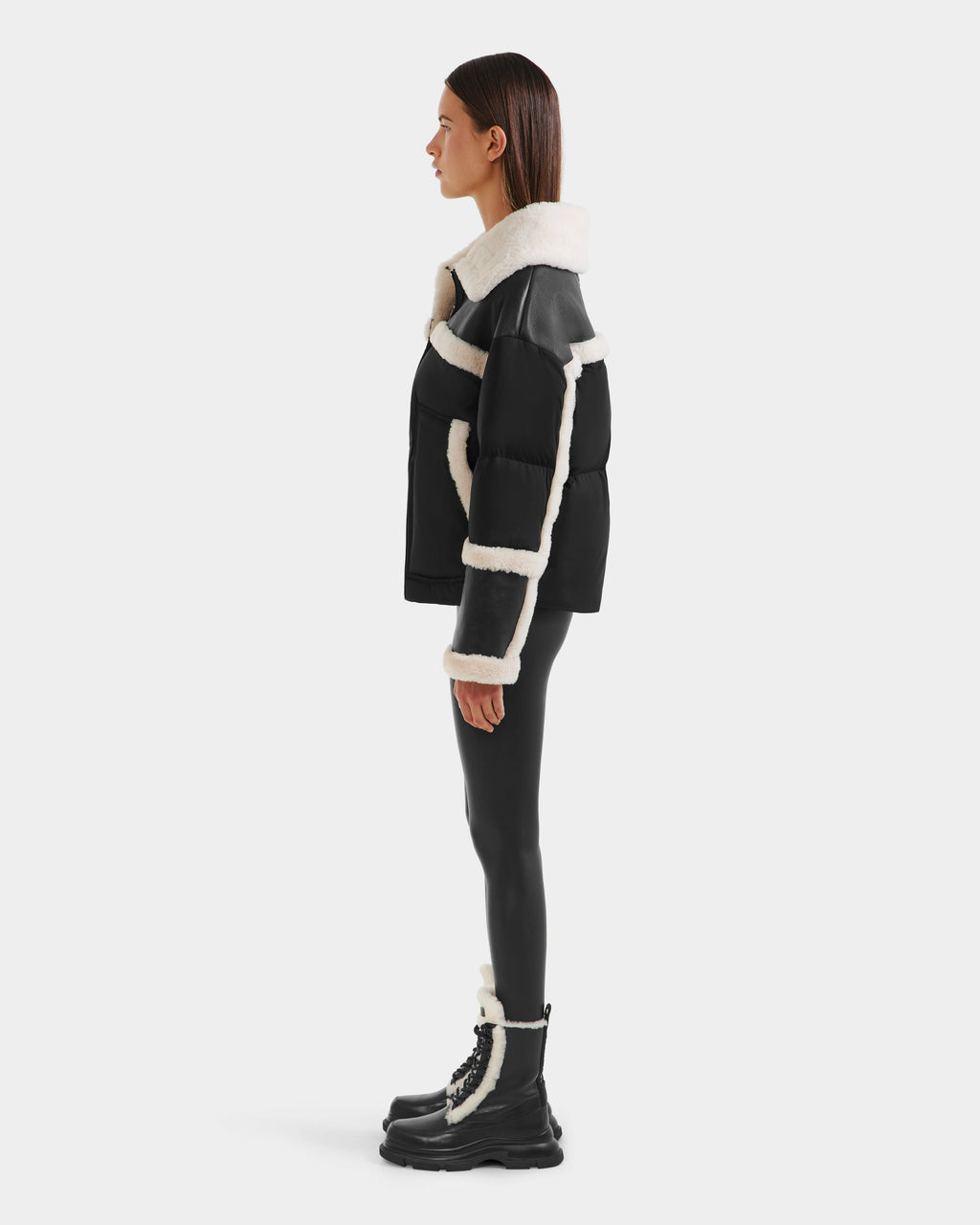 A person in profile showcases chic winter style wearing Rudsak’s SHIRLEY - 8125613 Black shearling jacket, glossy black leggings, and black lace-up boots with white trim, all set against a plain white background.