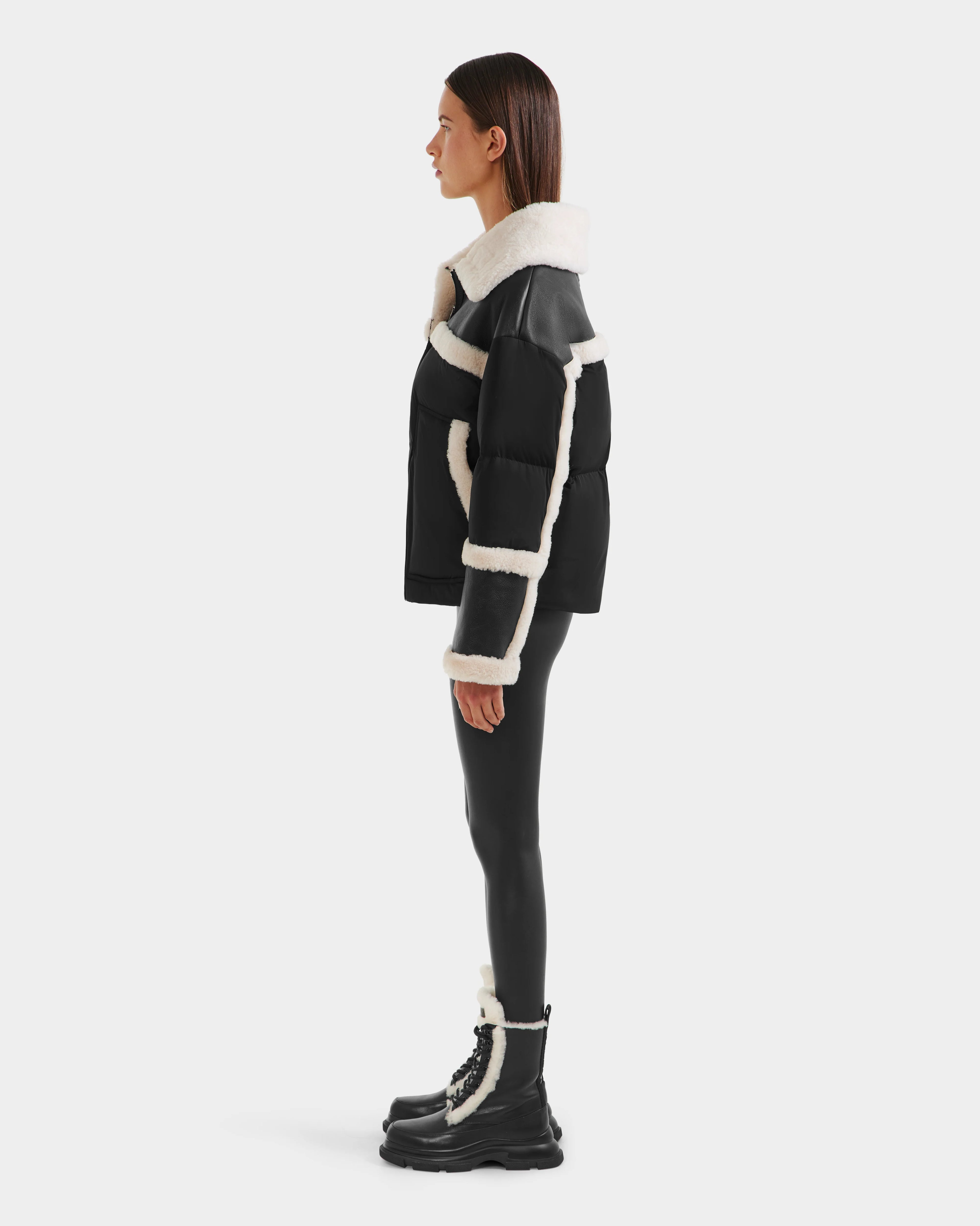 A person in profile showcases chic winter style wearing Rudsak’s SHIRLEY - 8125613 Black shearling jacket, glossy black leggings, and black lace-up boots with white trim, all set against a plain white background.