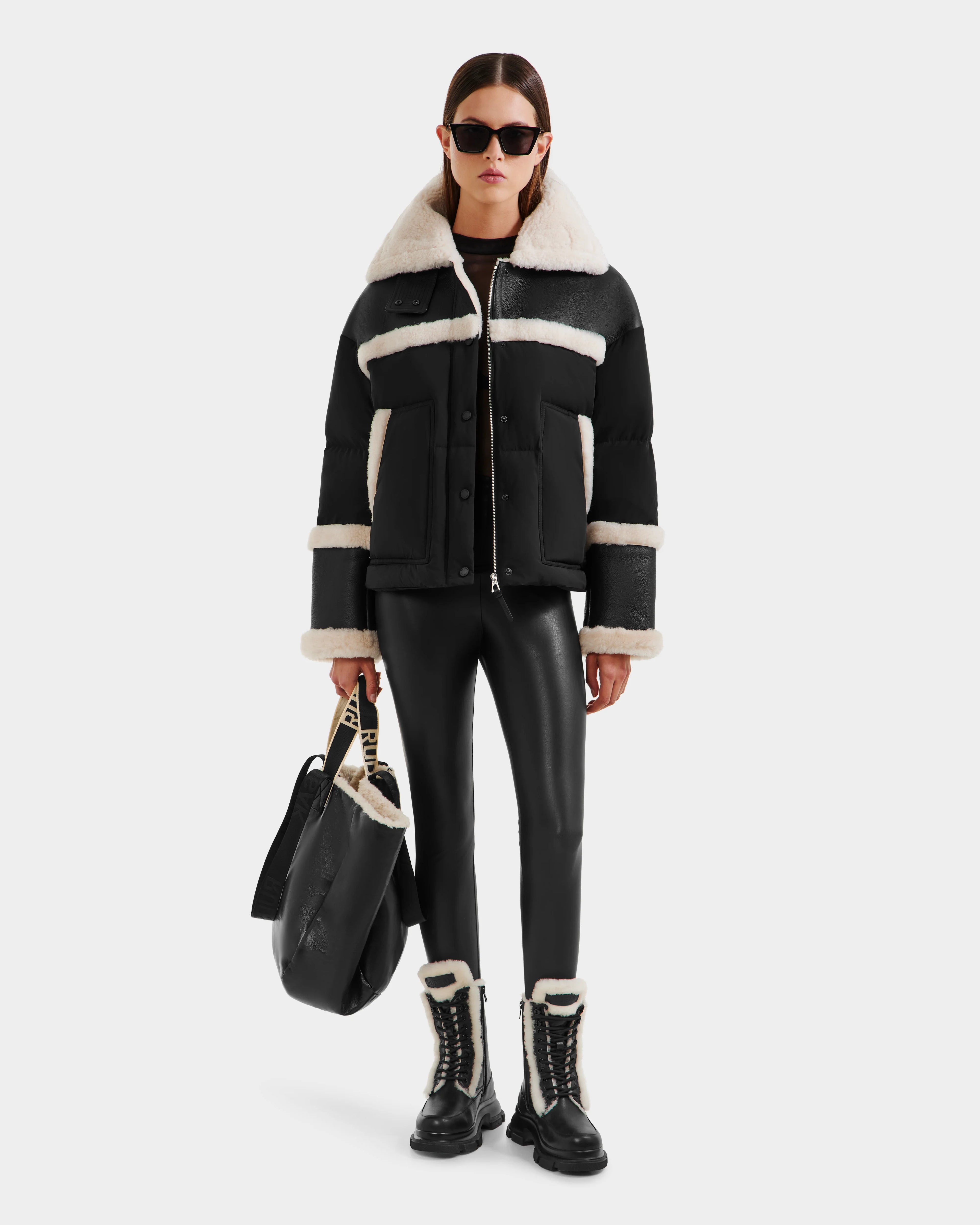 A woman models Rudsaks SHIRLEY - 8125613 Black jacket with black sunglasses, leggings, matching black boots with white trim, and a large black handbag, standing against a plain white background.