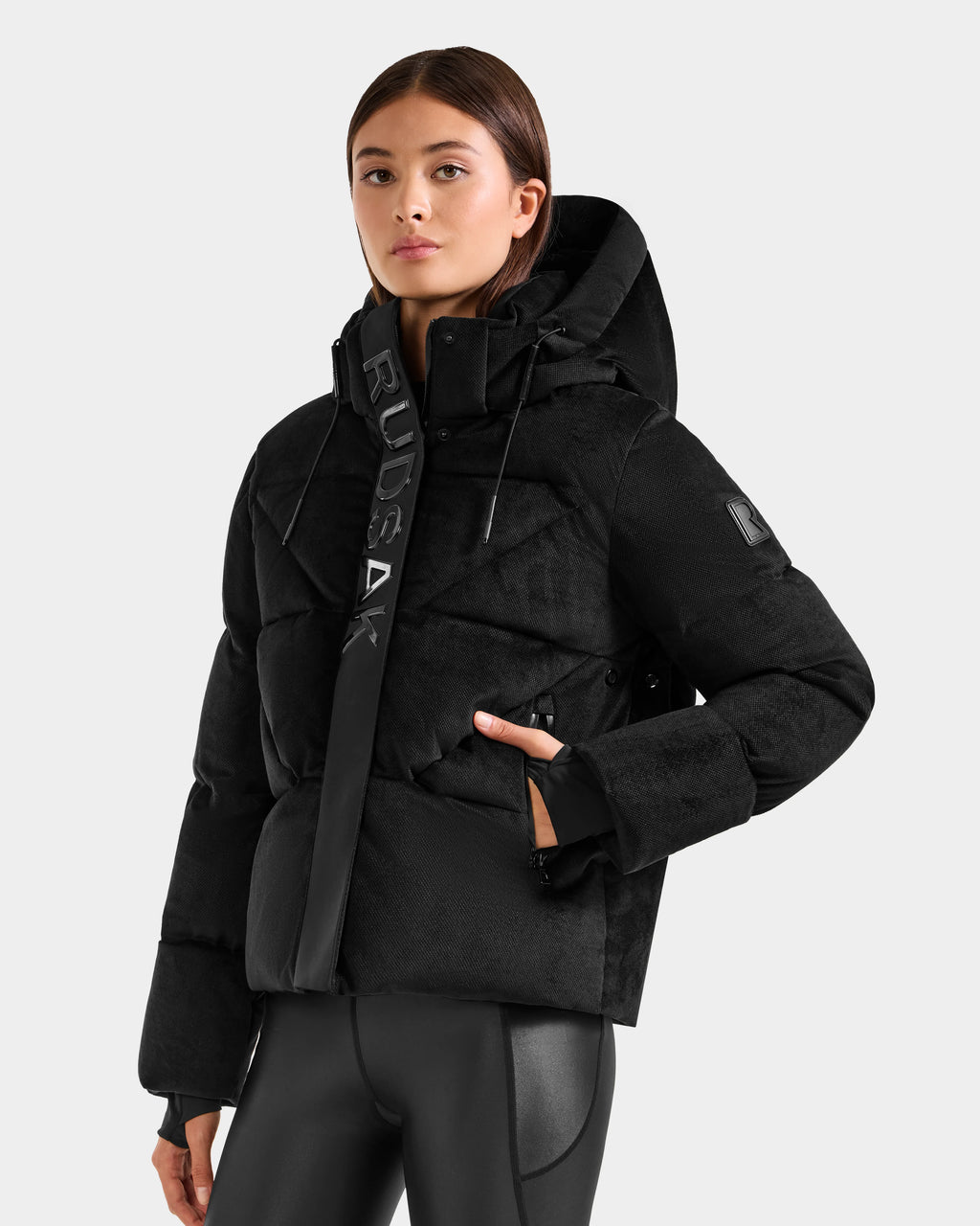 Someone wearing the Rudsak STEFANIE V - L125584 Black down puffer jacket with "RUDSAK" on the front collar stands against a light gray background. The winter coat features a high collar and zippered pockets.