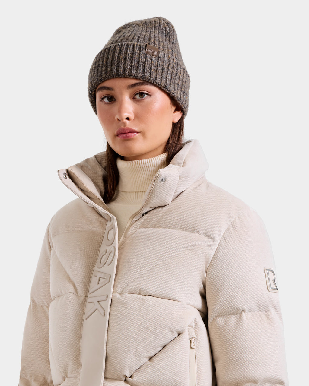 A woman wears the Rudsak STEFANIE V - L125584 Vanilla down puffer jacket with a high collar and light beige turtleneck, paired with a ribbed gray knit beanie—ideal for any winter wardrobe.