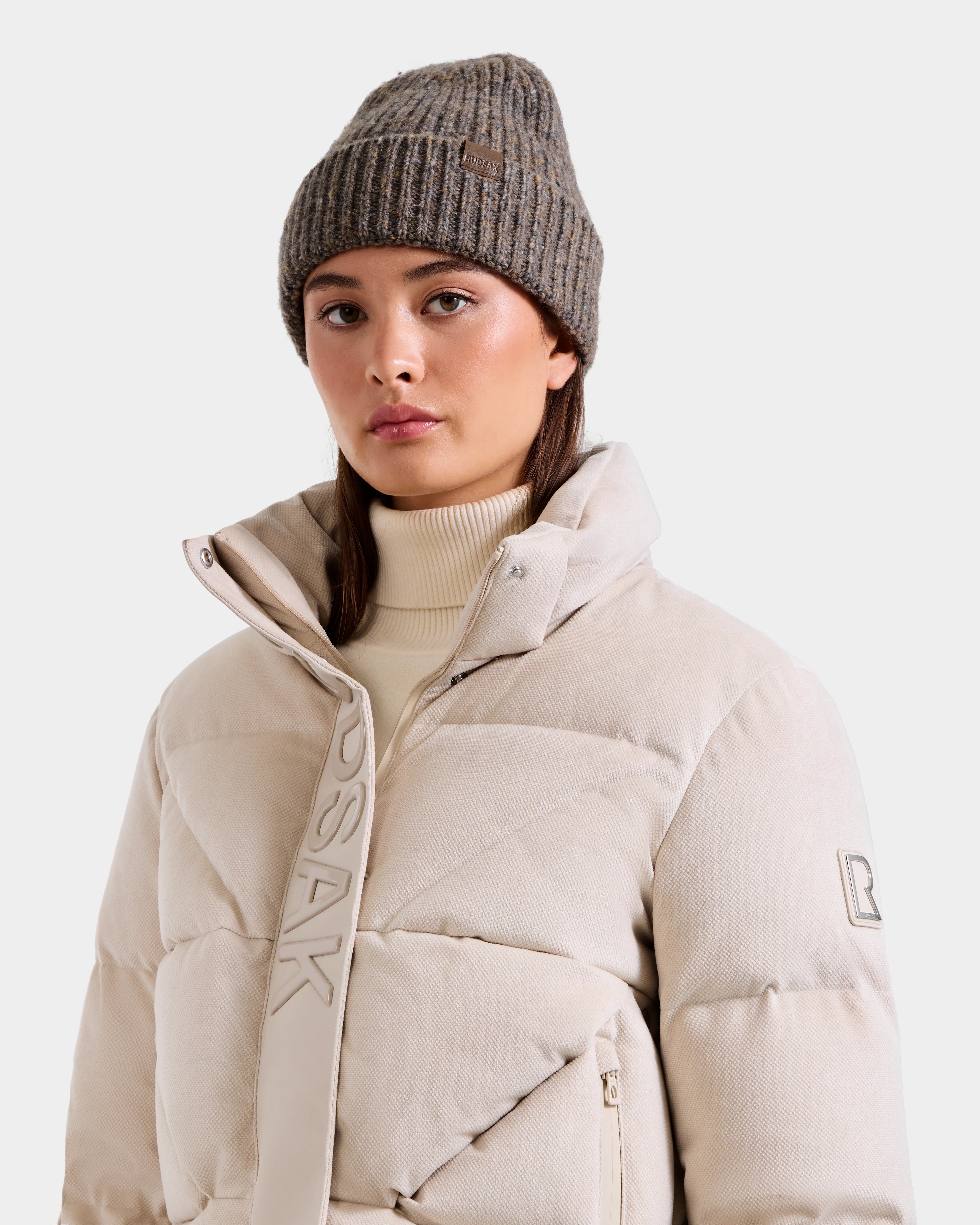 A woman wears the Rudsak STEFANIE V - L125584 Vanilla down puffer jacket with a high collar and light beige turtleneck, paired with a ribbed gray knit beanie—ideal for any winter wardrobe.